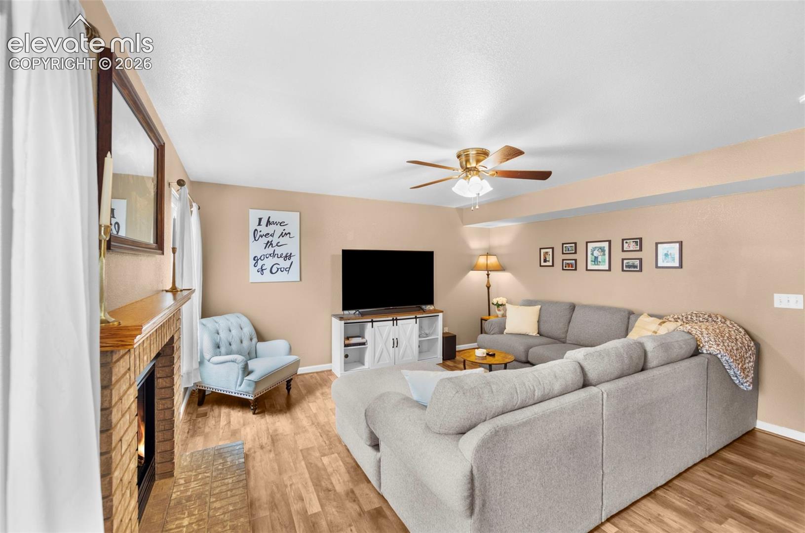 Living area featuring wood-finish flooring, a brick fireplace with a wood mantel, a ceiling fan with integrated lighting, and neutral wall tones