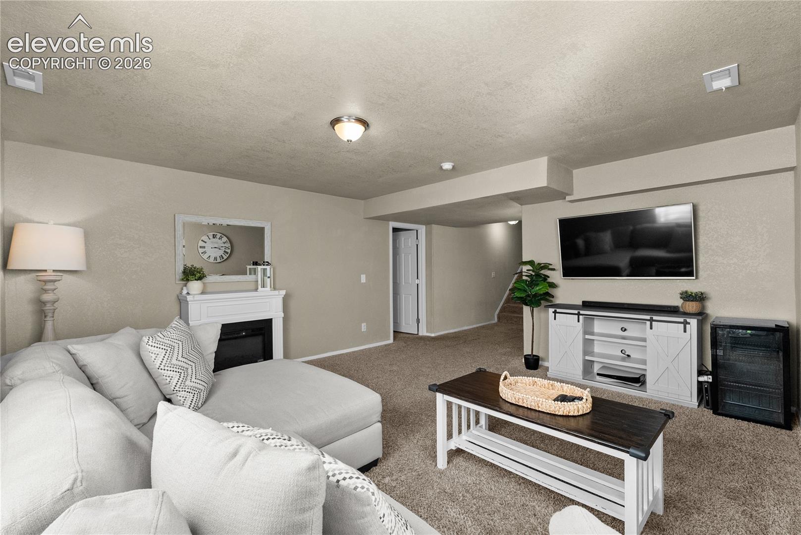Carpeted flooring, recessed ceiling lighting, wall-mounted television, built-in shelving, and a white fireplace mantel