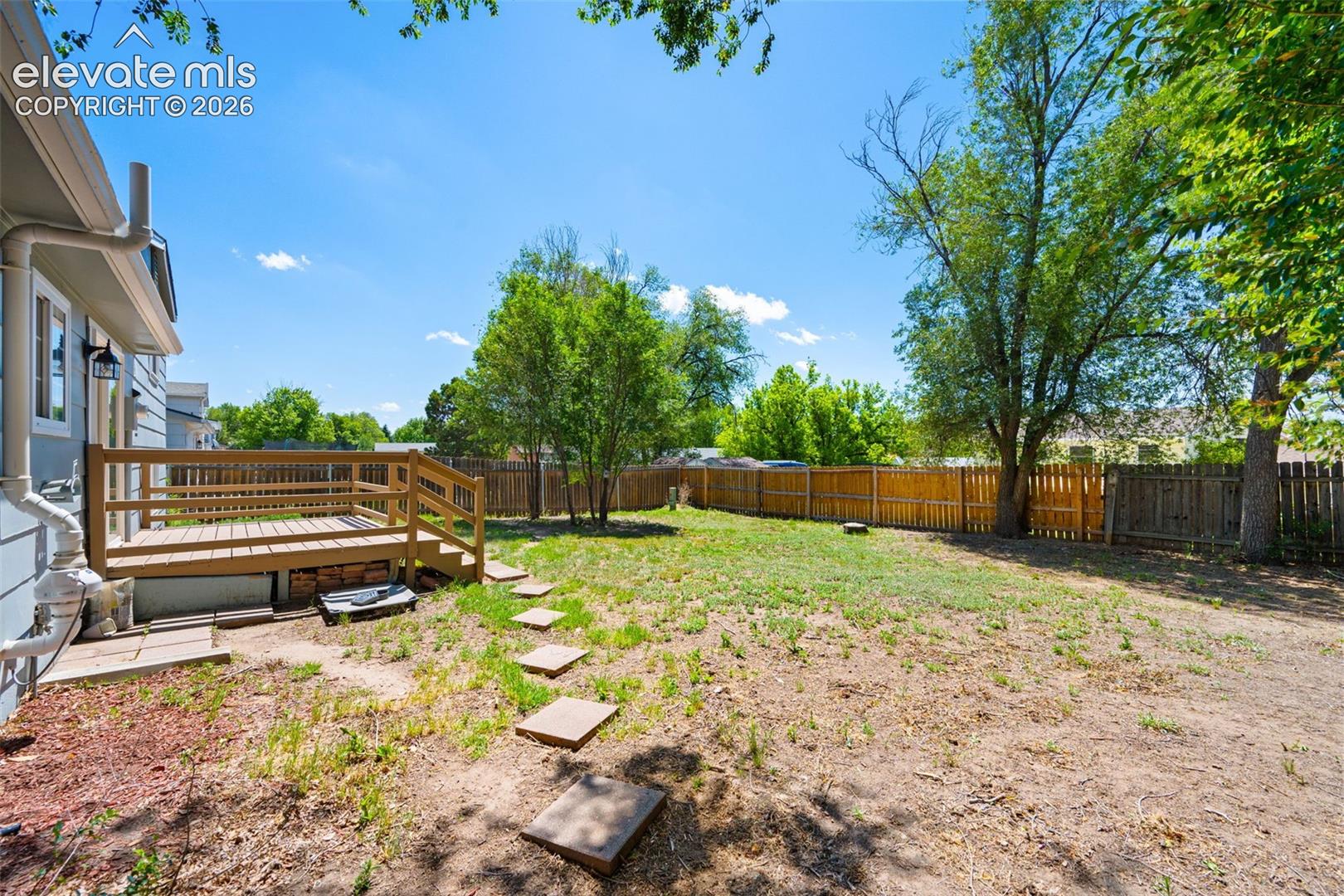 Wood deck with railing, fenced yard, mature trees, and stone stepping path