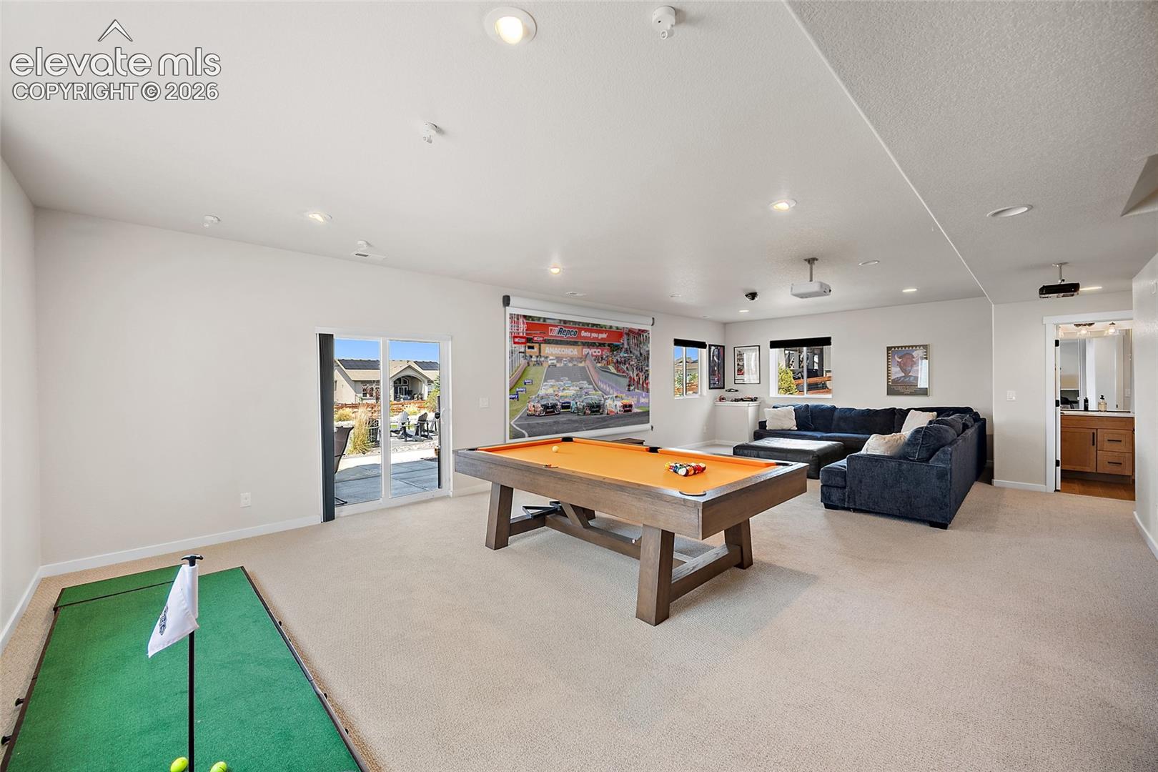 Spacious recreation room featuring recessed lighting, a sliding glass patio door, carpeted flooring, and a dedicated media wall with a projector screen