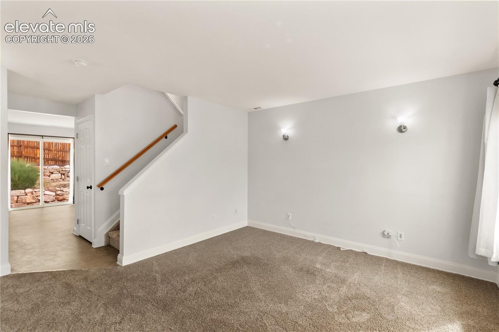 Neutral-toned carpeted flooring, wall-mounted light fixtures, staircase with a wood handrail, and glass sliding door access to exterior grounds