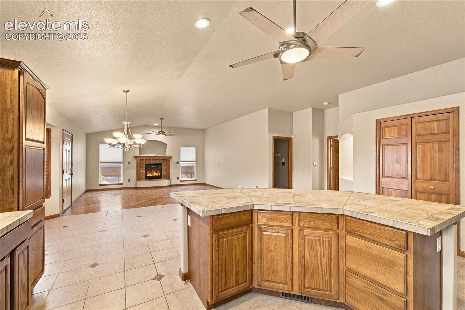 Kitchen island with tile countertop and wood-finish cabinetry, vaulted ceilings, fireplace with stone-finish surround, wood-finish flooring, and multi-light chandelier