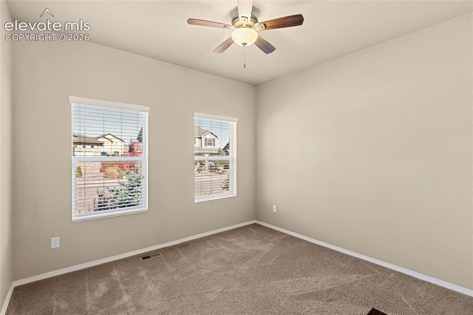 Neutral-toned interior space featuring carpeting, two windows with horizontal blinds, and a ceiling fan with integrated lighting