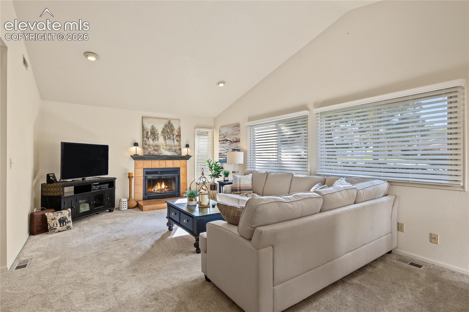 Vaulted ceiling, recessed lighting, gas fireplace with tile surround, carpeted flooring, and multiple windows with blinds