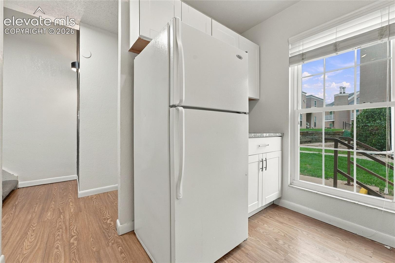 Kitchen featuring white shaker-style cabinetry, wood-finish flooring, a multi-pane window, and integrated refrigerator