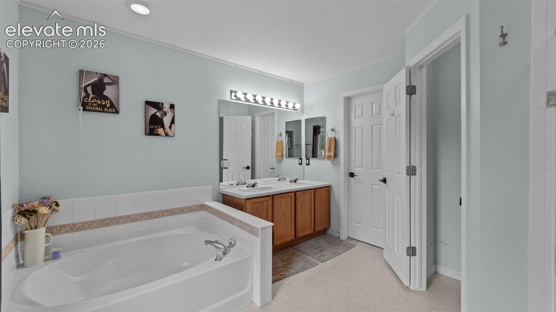Built-in soaking tub with tile surround, double-sink vanity with wood cabinetry, large vanity mirror, multi-bulb light fixture, and six-panel interior doors