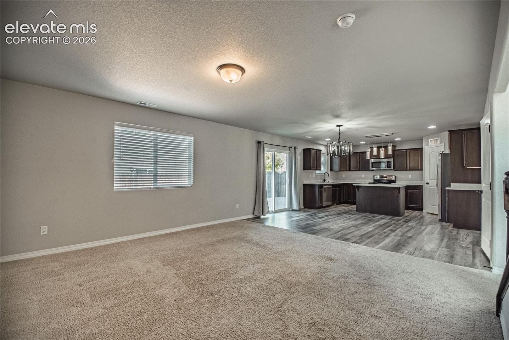Expansive living area with carpet flooring, window with blinds, and a built-in ceiling light