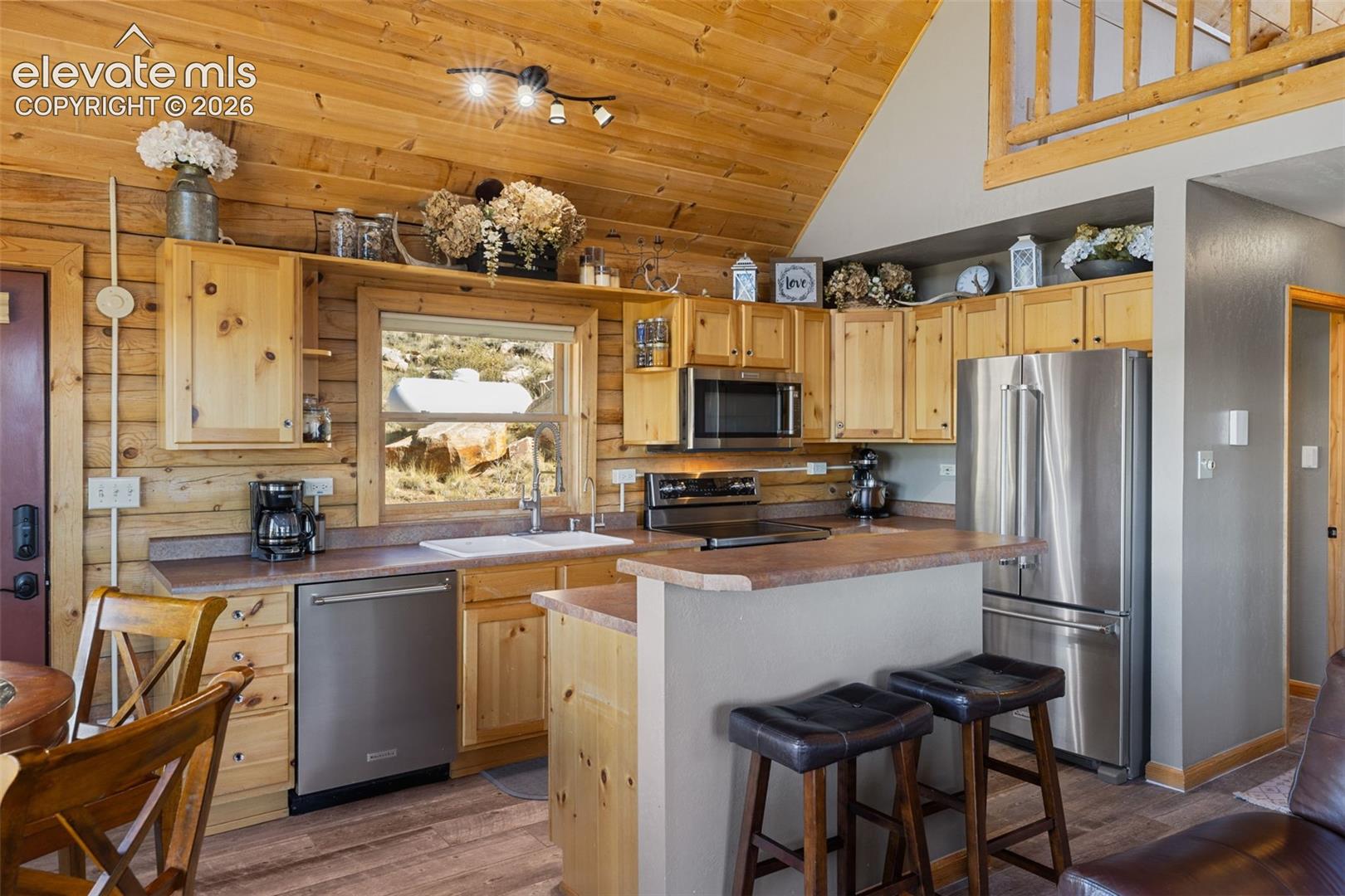 Vaulted wood-plank ceiling, natural log walls, stainless steel kitchen appliances, custom wood cabinetry, and a peninsula counter