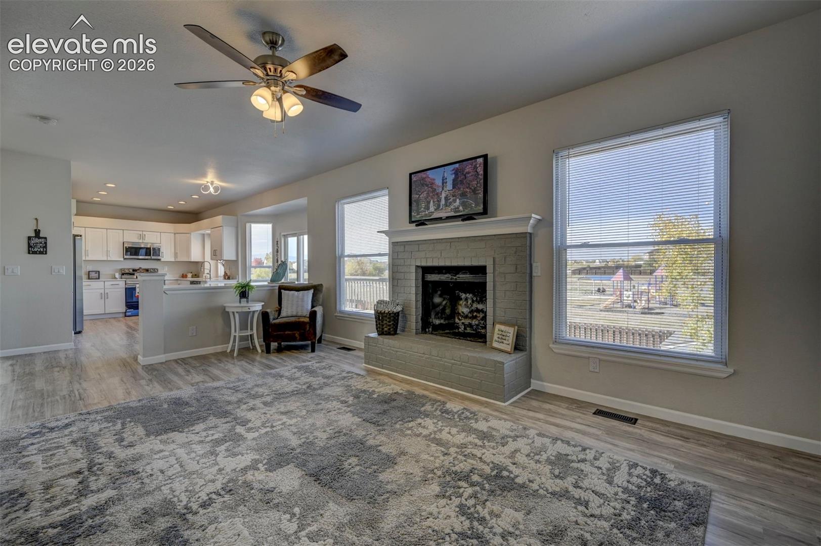 Living area featuring wood-finish flooring, a brick fireplace with a painted finish, a ceiling fan with integrated lighting, and large windows