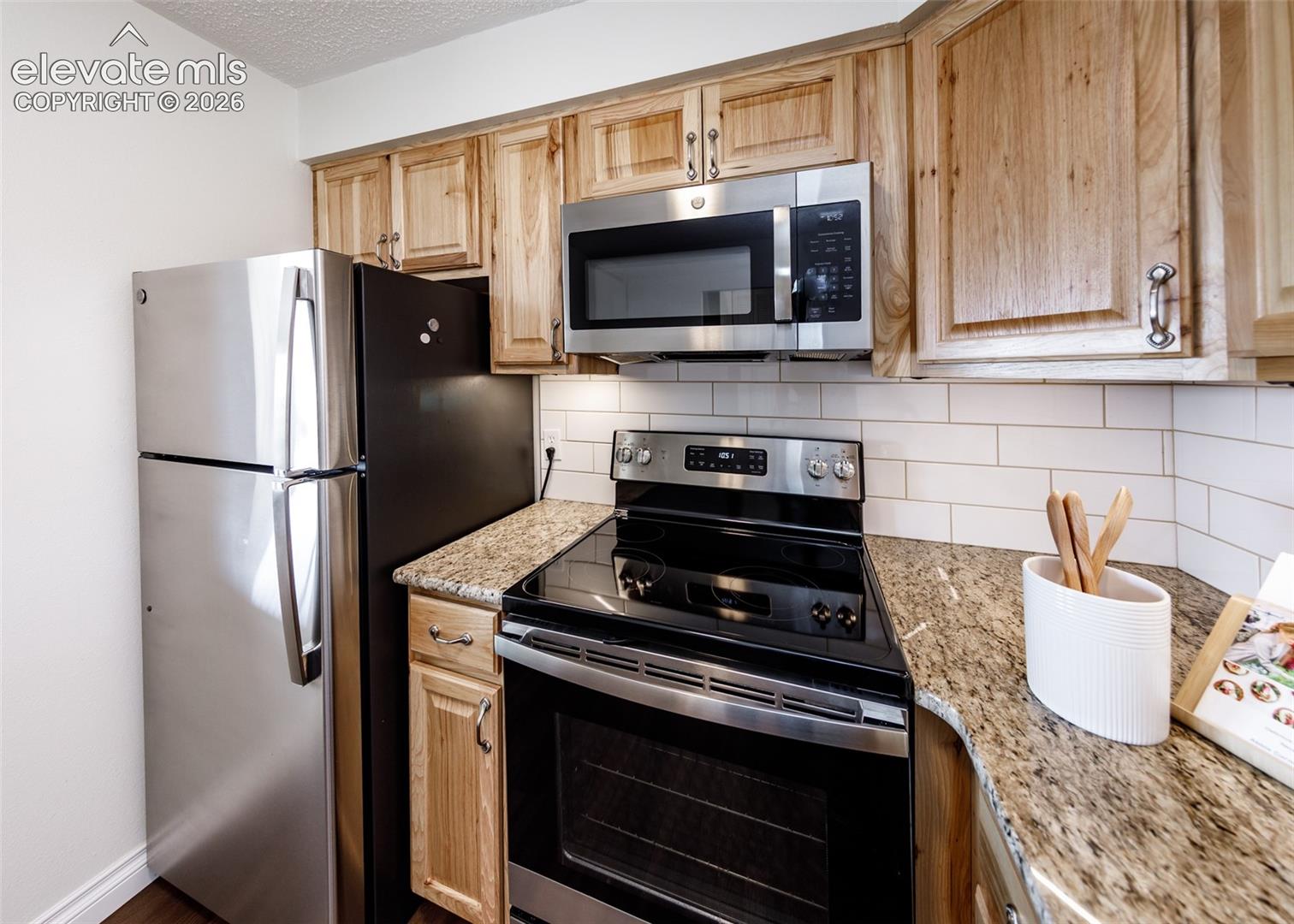 Kitchen featuring natural wood cabinetry, stone-surface countertops, subway tile backsplash, and stainless steel appliances including a built-in microwave and refrigerator