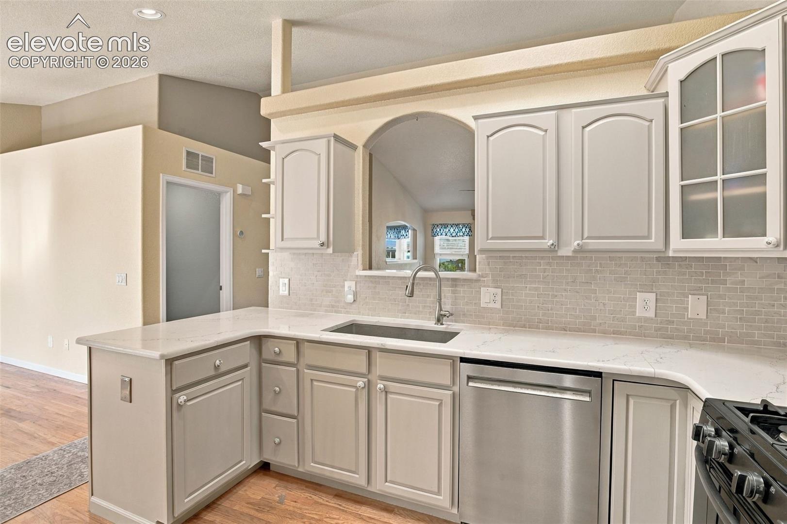 Kitchen featuring light gray shaker-style cabinetry, white stone-composite countertops, brick-patterned tile backsplash, stainless steel dishwasher, and a wide pass-through opening