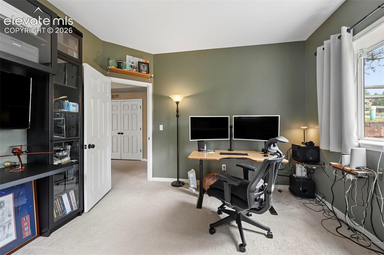 Window with outdoor scenery, light-colored carpeting, warm-toned wall paint, and a built-in shelving unit with glass-fronted cabinets