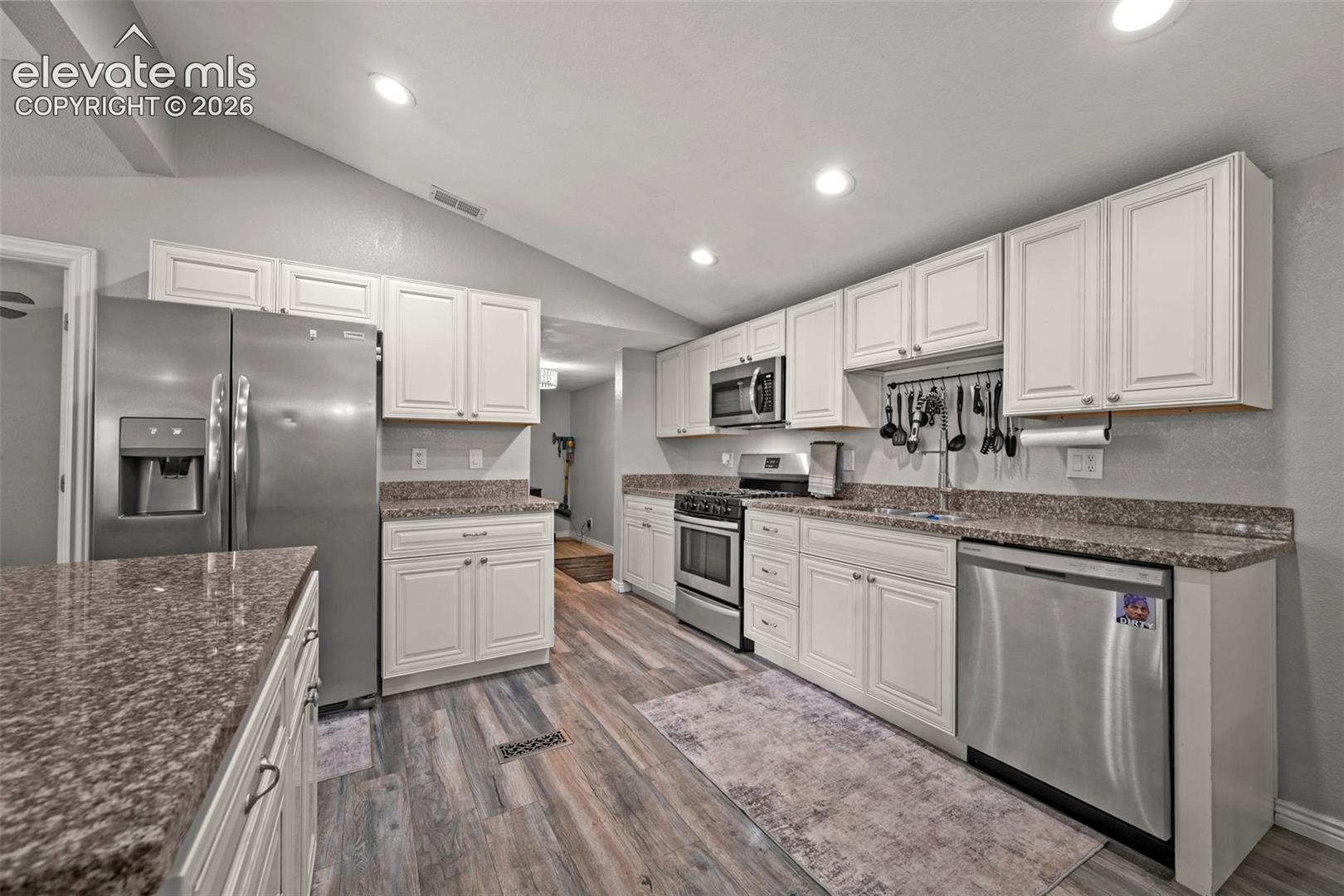Kitchen featuring white shaker-style cabinetry, stone-surface countertops, wood-finish flooring, vaulted ceilings with recessed lighting, and stainless steel appliances
