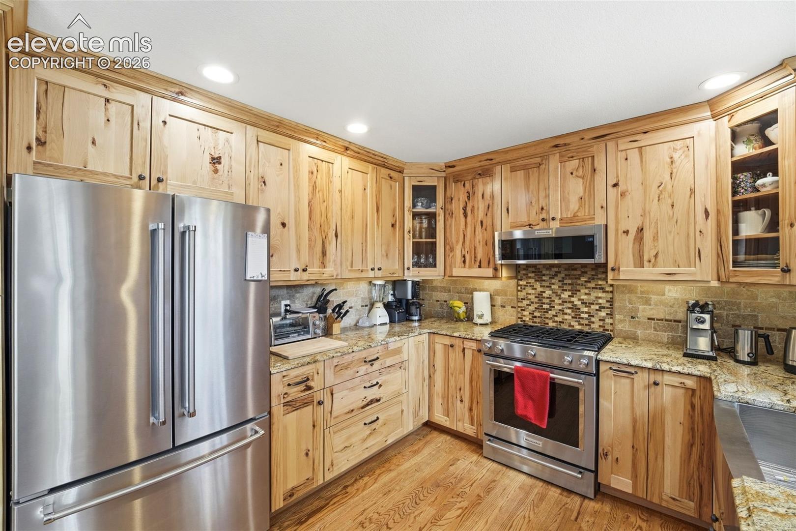 Kitchen featuring natural wood cabinetry, stainless steel appliances, granite countertops, a mosaic tile backsplash, and wood-finish flooring