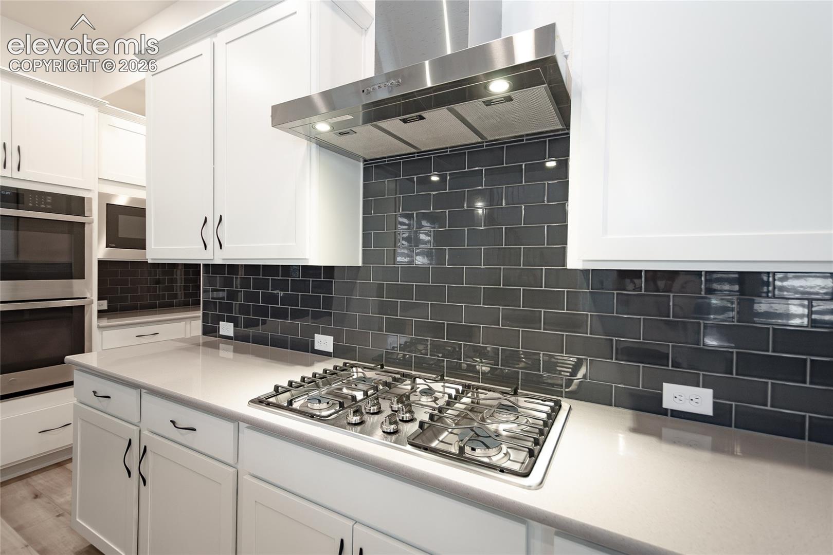 Kitchen featuring white shaker-style cabinetry, a dark subway tile backsplash, a stainless steel gas cooktop, a stainless steel range hood, and a light-toned countertop