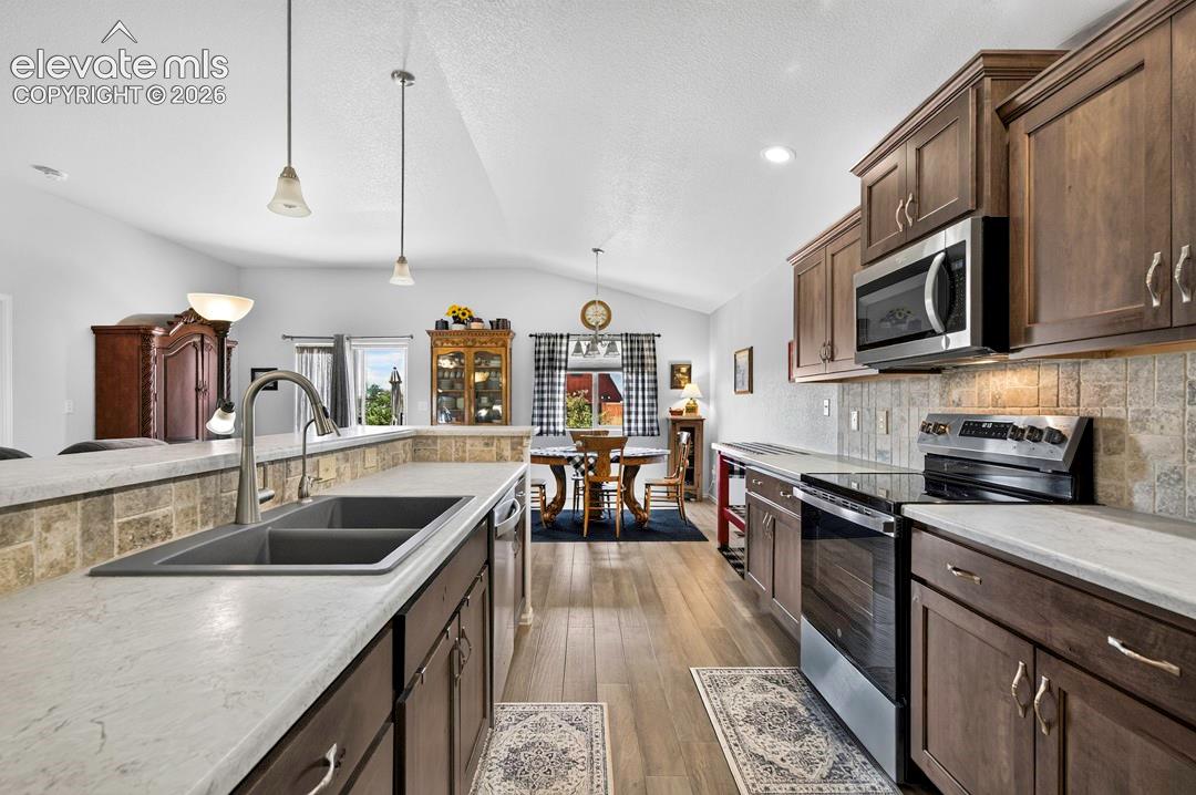 Open-concept kitchen featuring wood-finish flooring, dark wood cabinetry, a tiled backsplash, a stainless steel electric range, and an integrated double sink with a gooseneck faucet