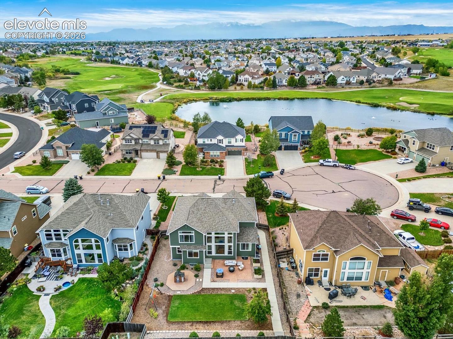 Aerial perspective featuring a residential property with a rear stone patio, built-in fire pit, and level lawn, set against a backdrop of a community pond and distant mountain range