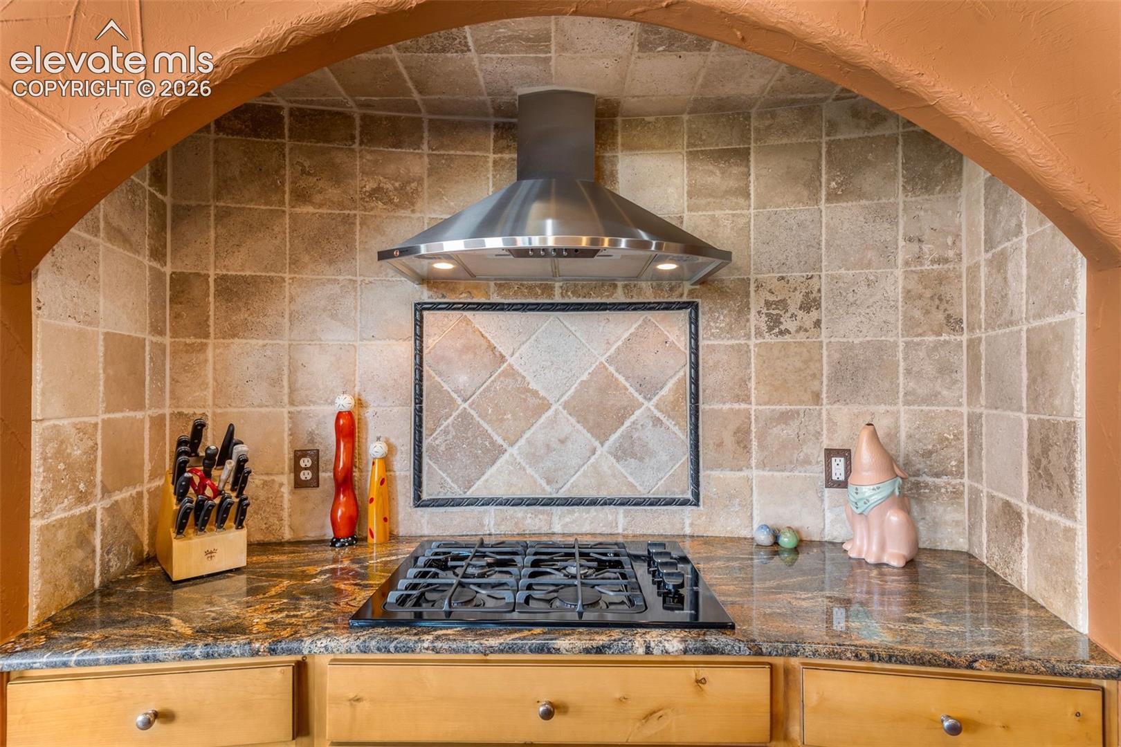 Arched stove alcove featuring a stone-tiled backsplash with decorative inlay, a stainless steel range hood, and a gas cooktop set into a stone countertop