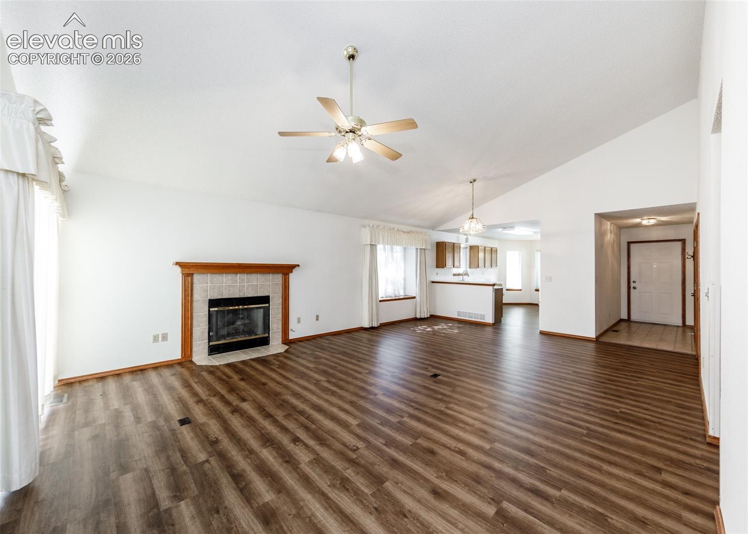 Vaulted ceiling with a ceiling fan, wood-finish flooring, tiled fireplace with a wood mantle, and a kitchen pass-through