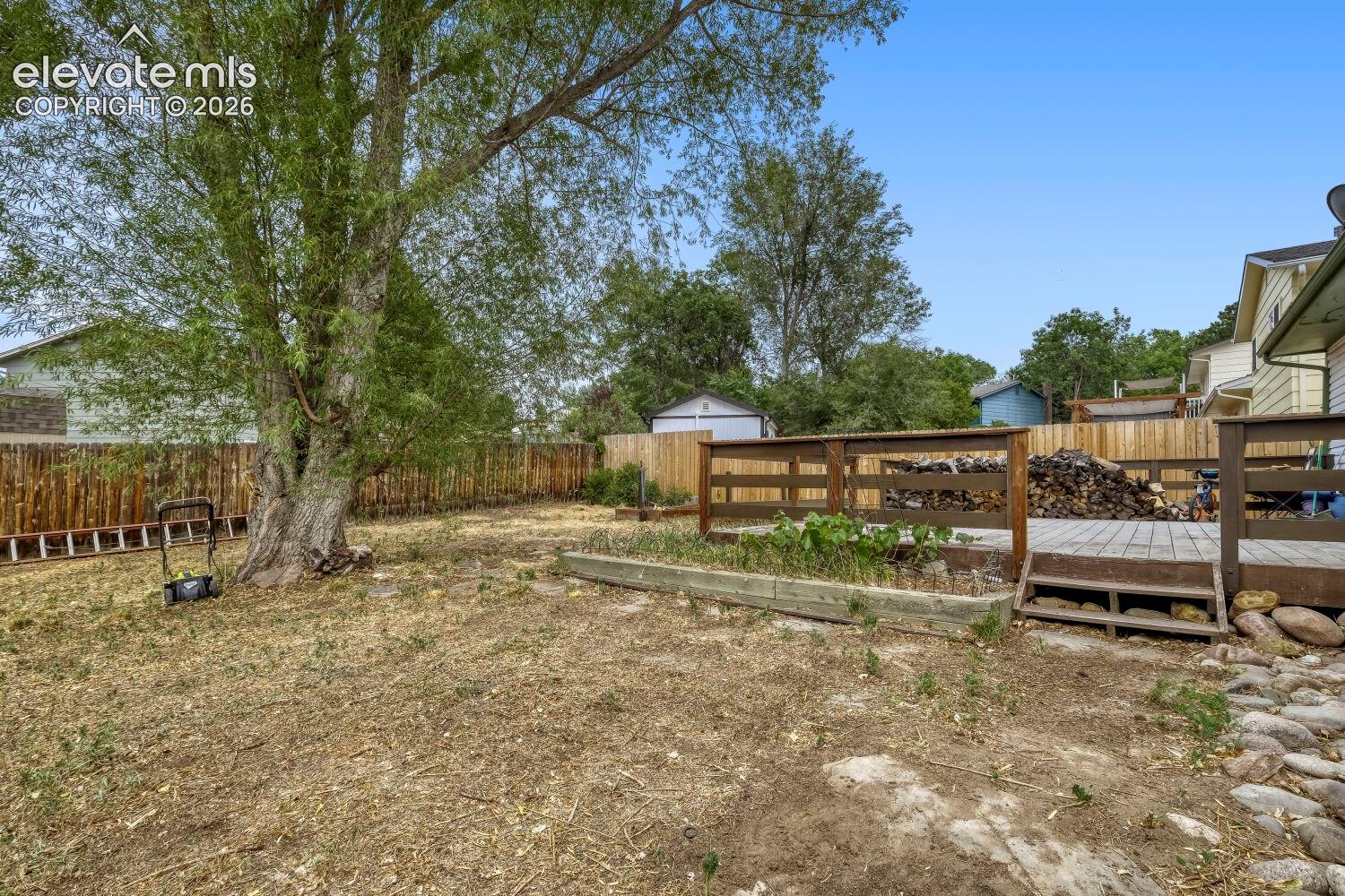 Spacious backyard featuring a large tree, wooden fencing, a raised garden bed, and a wooden deck with steps