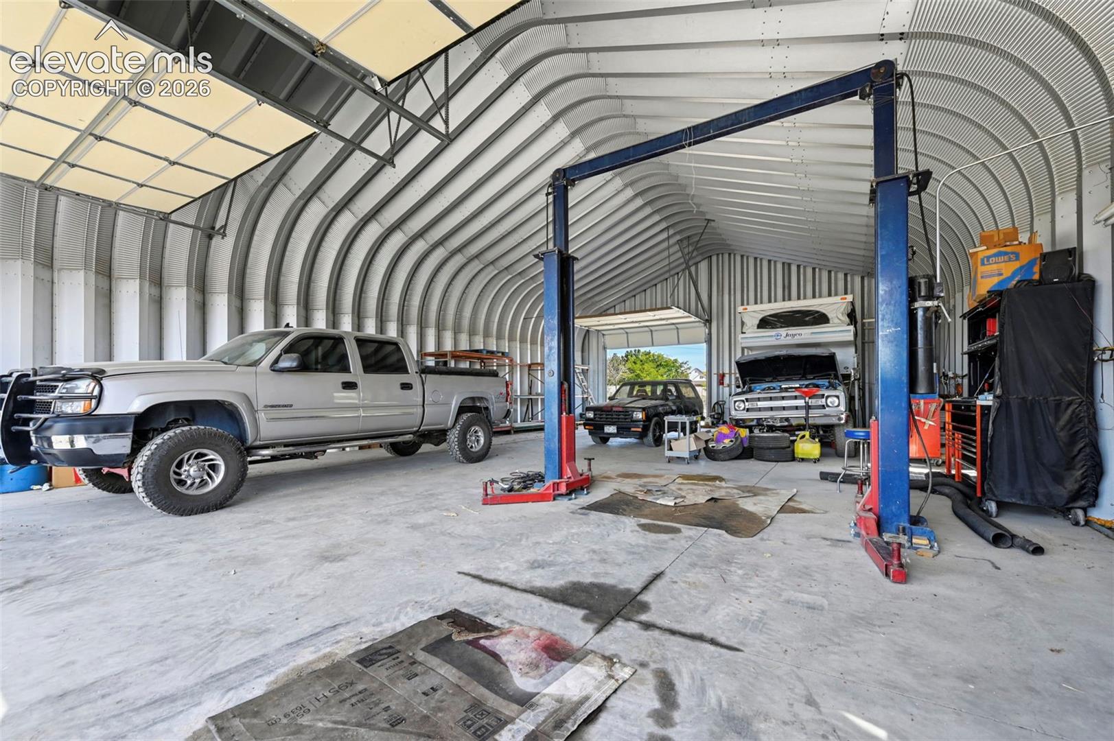 Quonset-style structure featuring a high-clearance vaulted ceiling, corrugated metal walls, concrete flooring, overhead rolling door, and an integrated hydraulic vehicle lift