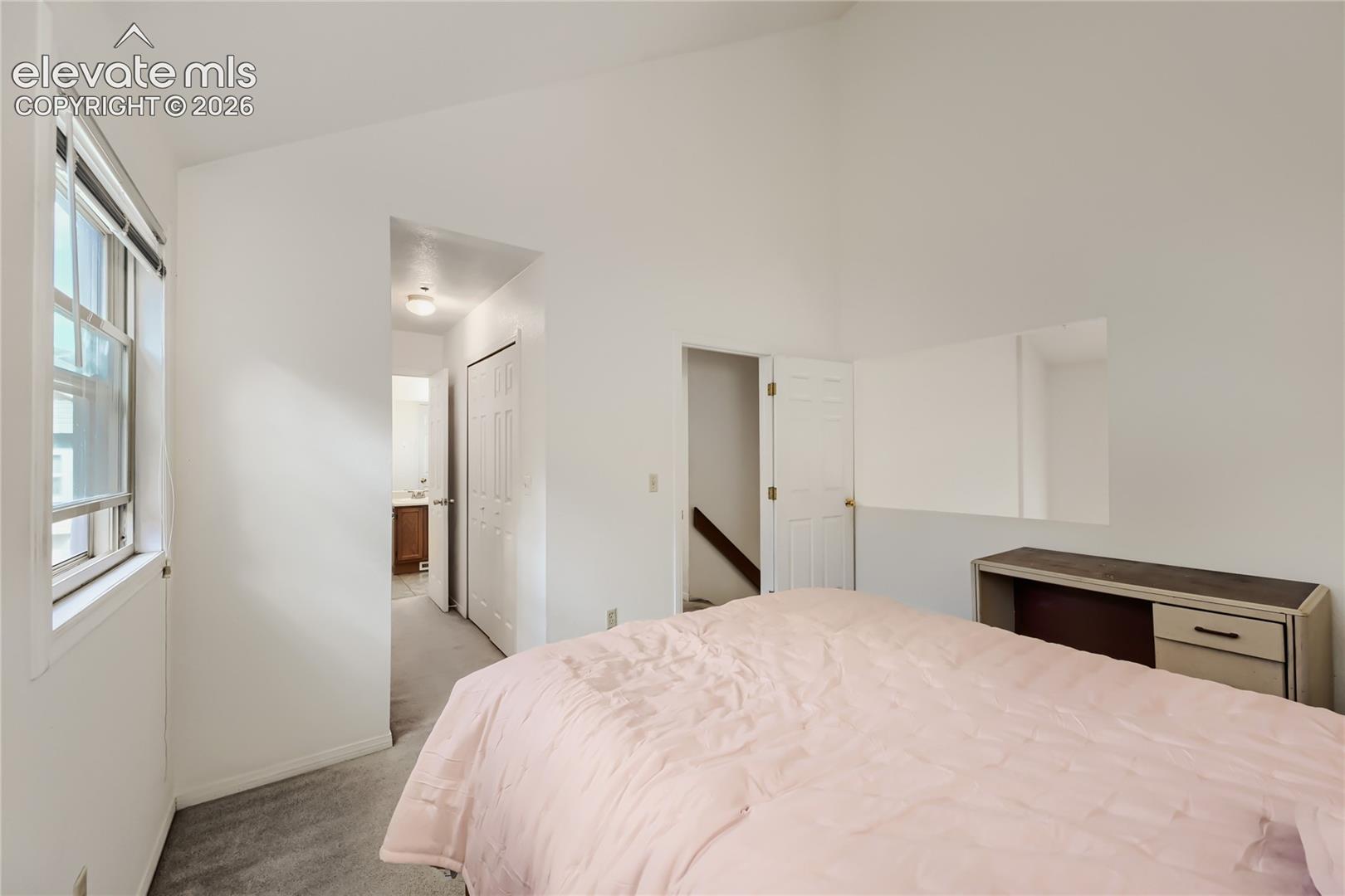 Vaulted ceiling, white walls, carpet flooring, a window with horizontal blinds, and a built-in shelf