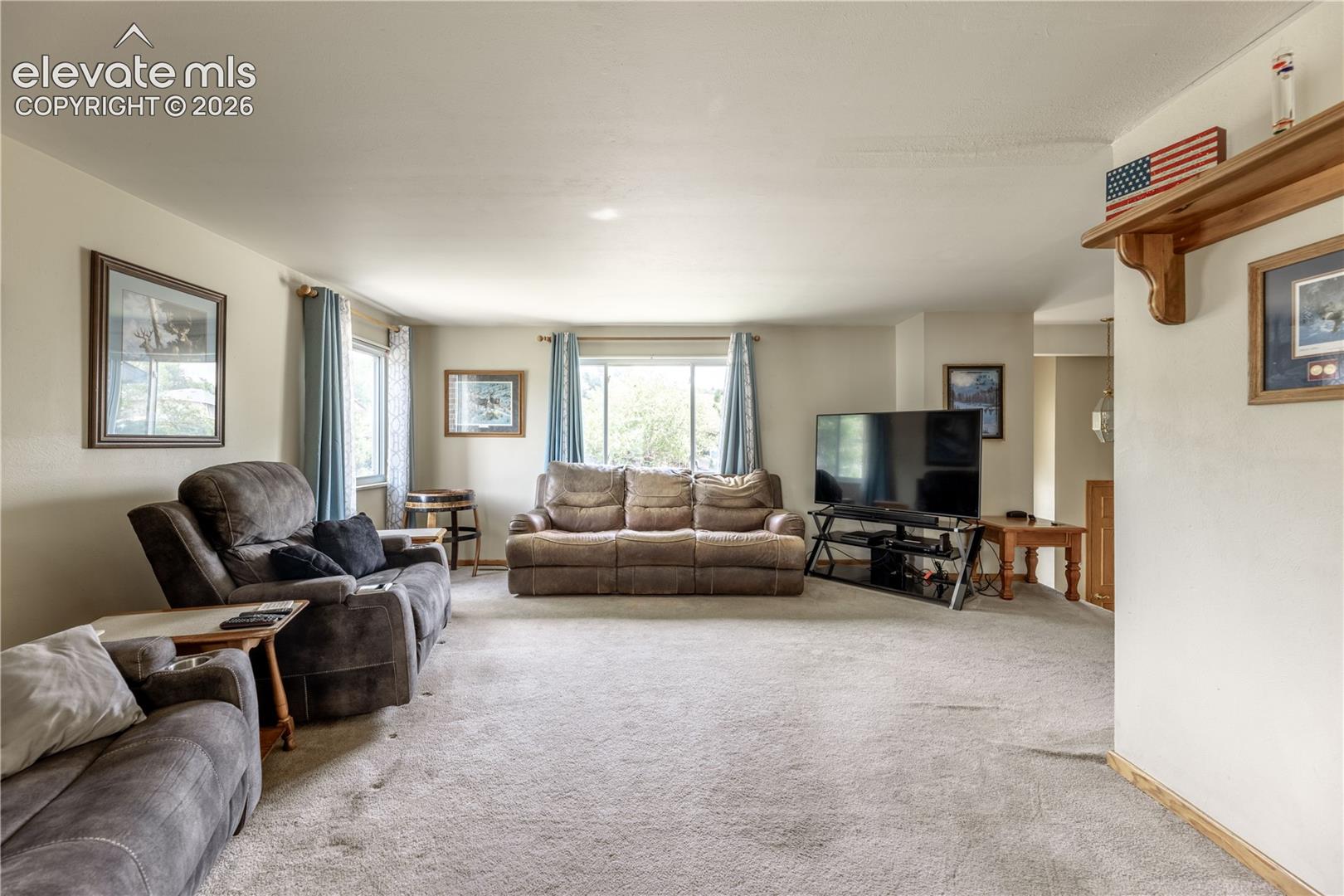 Neutral-toned living space featuring wall-to-wall carpeting, multiple windows providing natural light, and a built-in wooden wall shelf