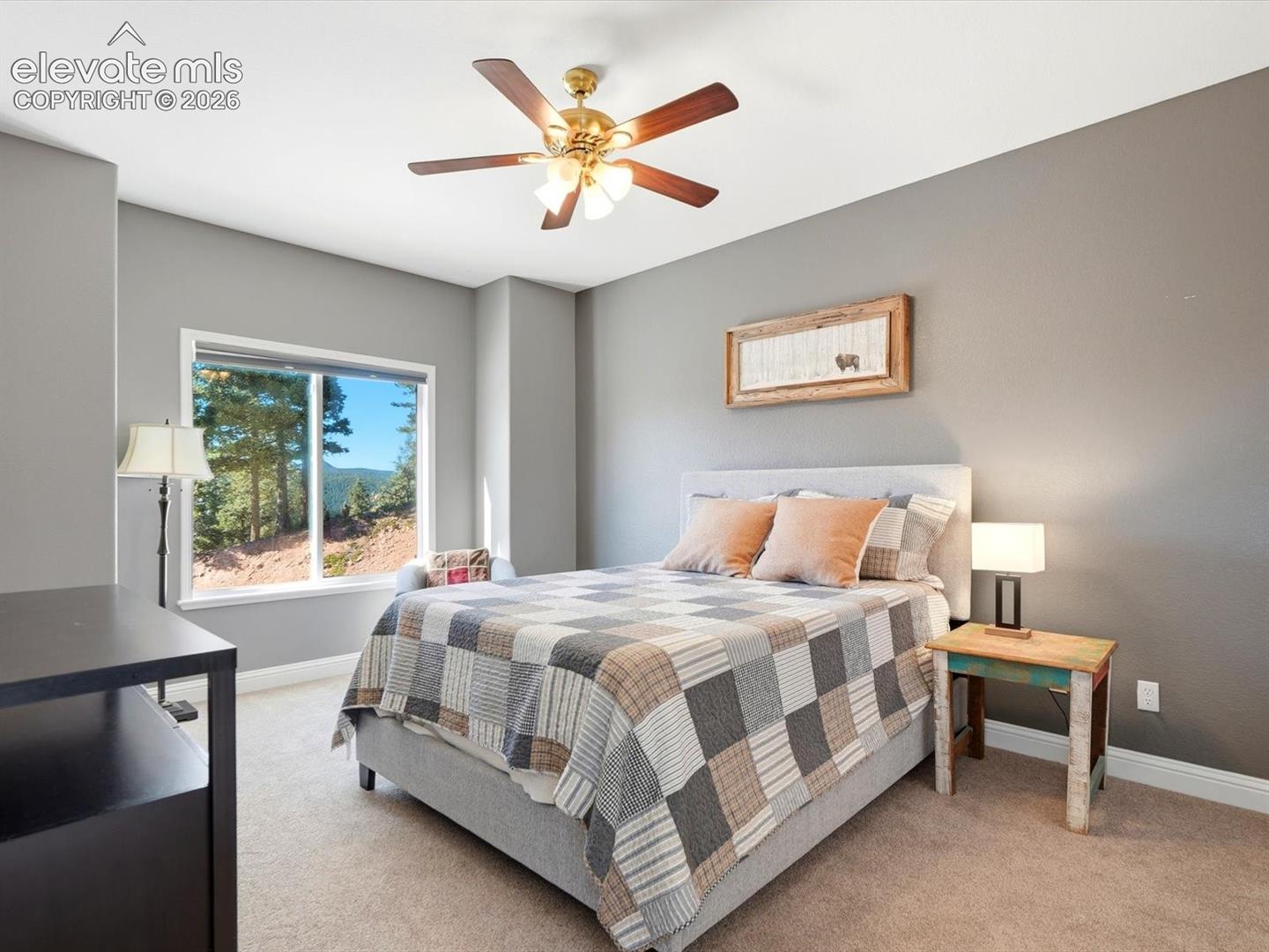Main floor secondary Bedroom featuring neutral wall tones, neutral carpeting, a multi-blade ceiling fan with integrated lighting, and a window with exterior forest scenery