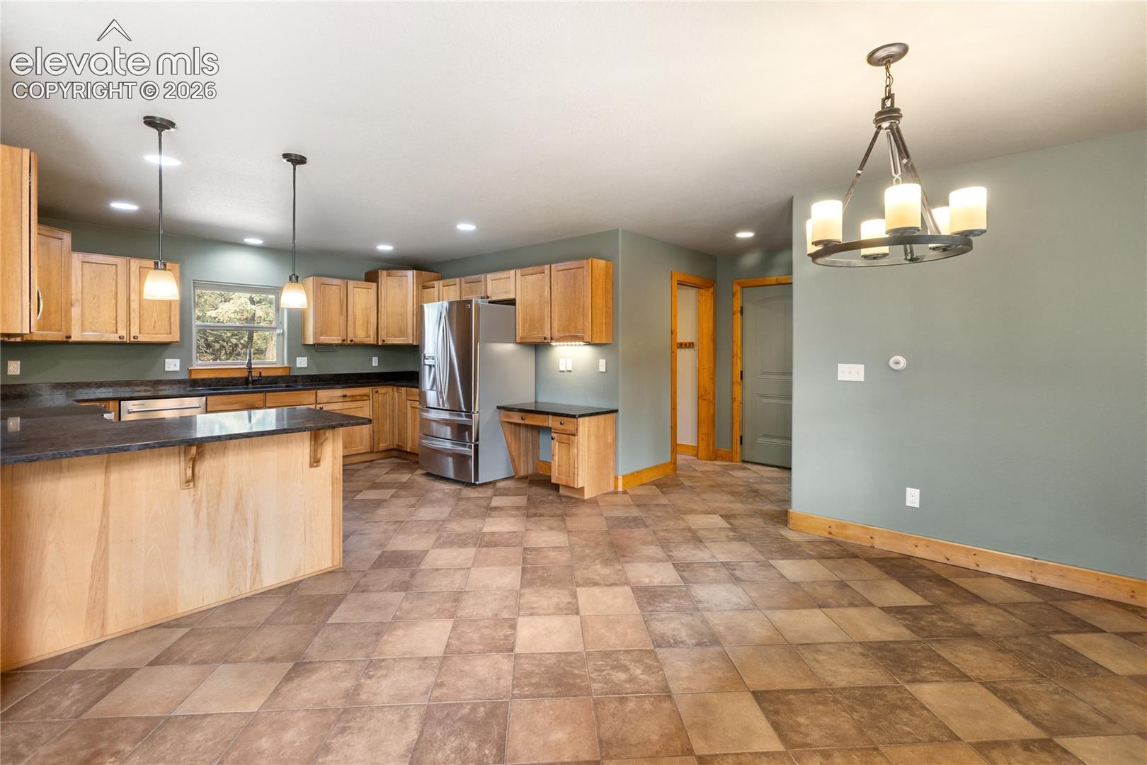 Kitchen featuring light wood cabinetry, dark stone countertops, stainless steel refrigerator, recessed lighting, and a hanging metal chandelier