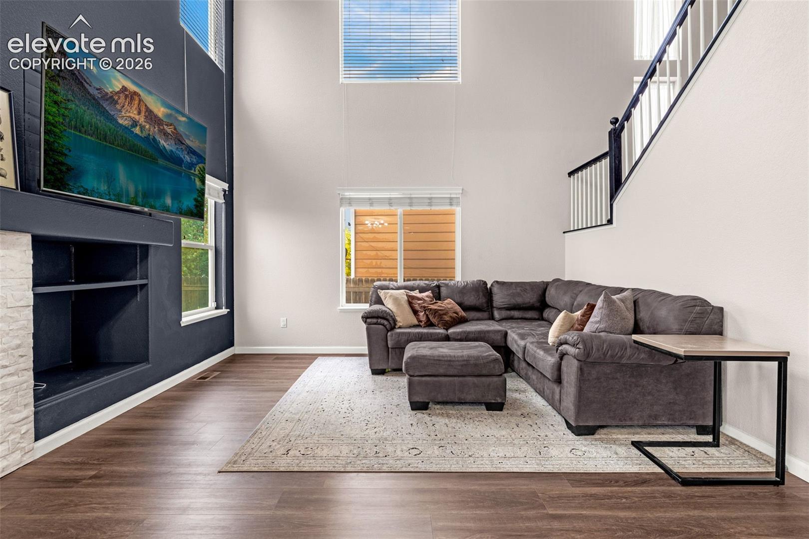 Double-height ceiling, floor-to-ceiling accent wall, fireplace with white stone-finish surround, wood-finish flooring, and staircase with black-and-white railings