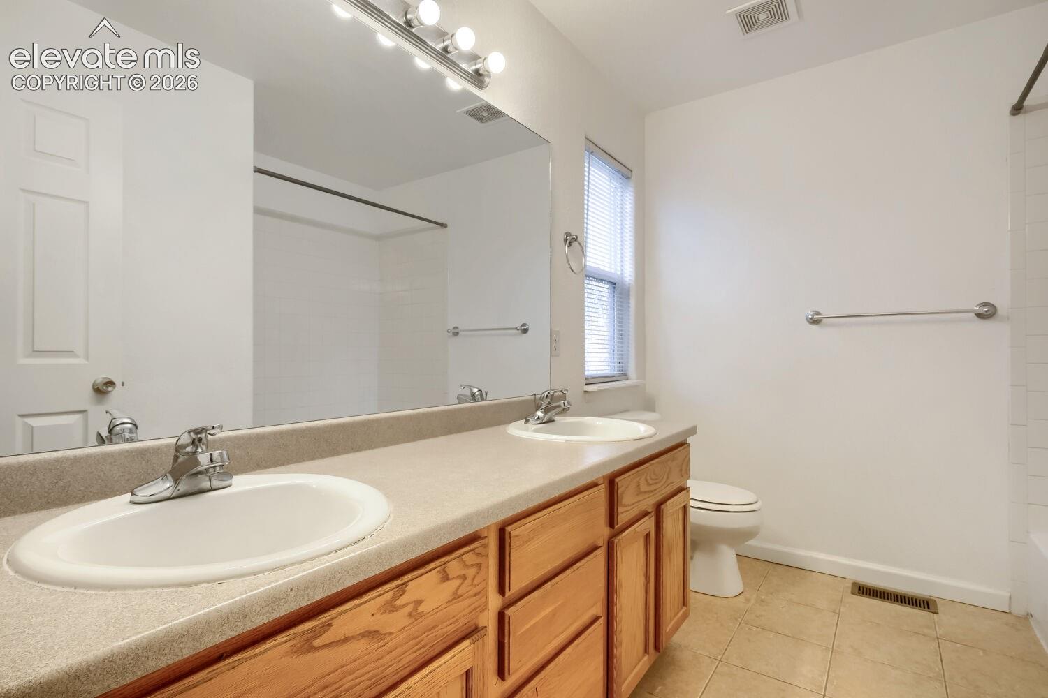 Bathroom featuring a dual-sink vanity with a solid-surface countertop, wood cabinetry, a large wall-mounted mirror, and tile flooring