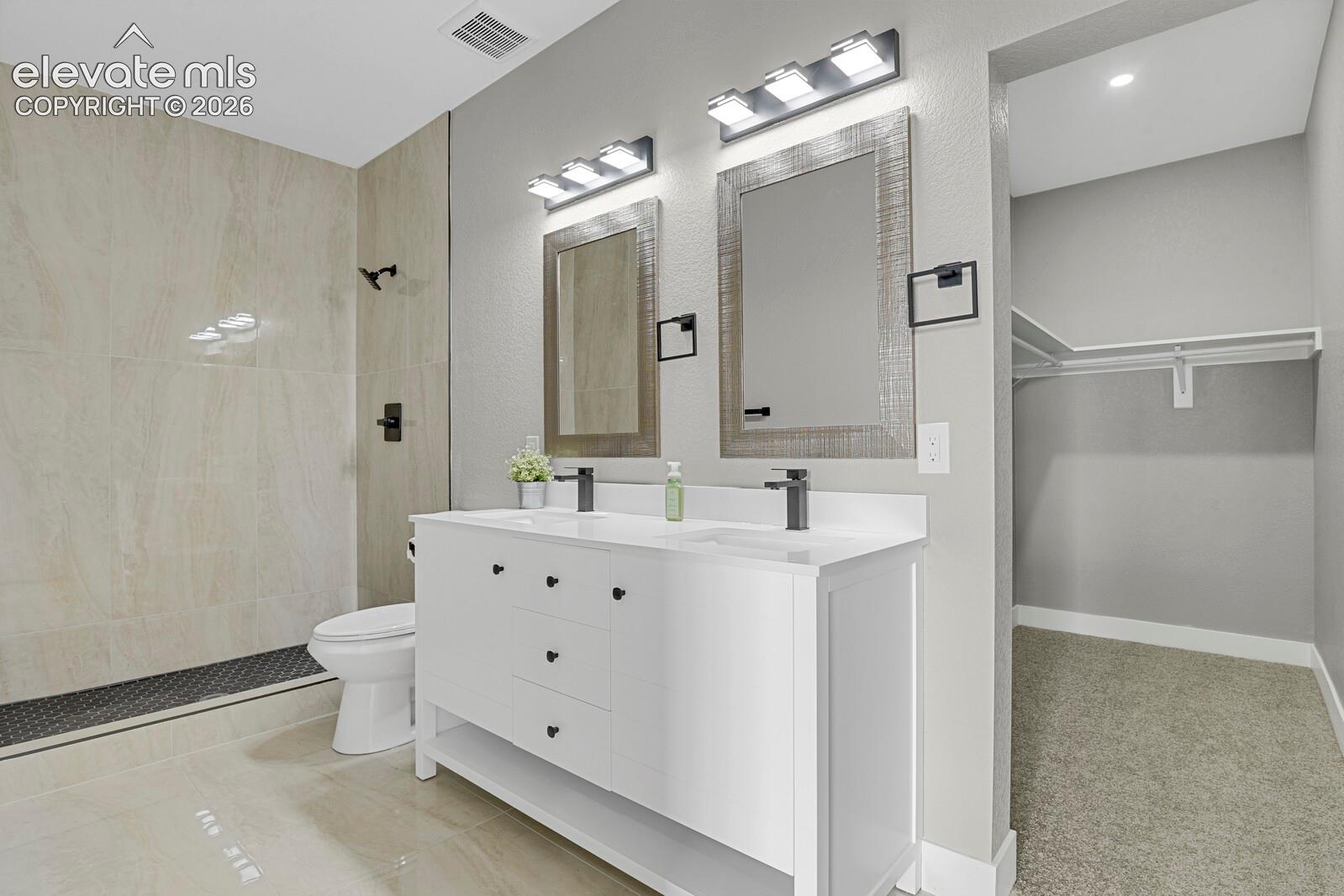 Dual-sink vanity with a white countertop and dark hardware, walk-in shower featuring tile surrounds, wall-mounted light fixtures, and a walk-in closet with built-in shelving