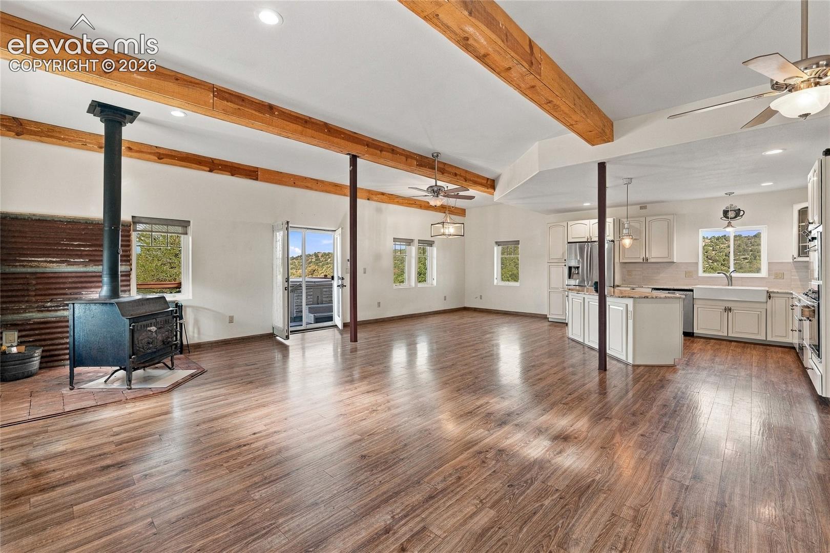 Open-concept living space featuring wood-finish flooring, exposed wood ceiling beams, a freestanding wood-burning stove with a corrugated metal accent wall, and a kitchen equipped with white cabinetry and a farmhouse-style sink