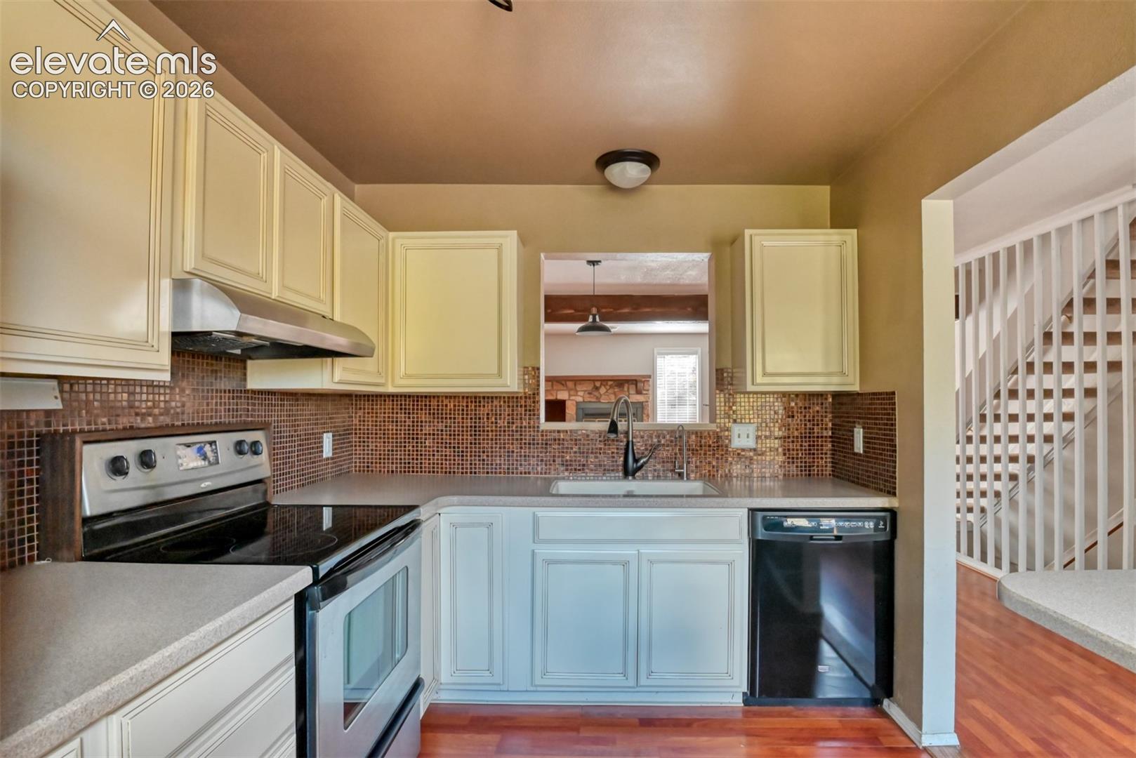 Kitchen featuring light-toned cabinetry, a mosaic tile backsplash, a stainless steel range hood, and a black dishwasher