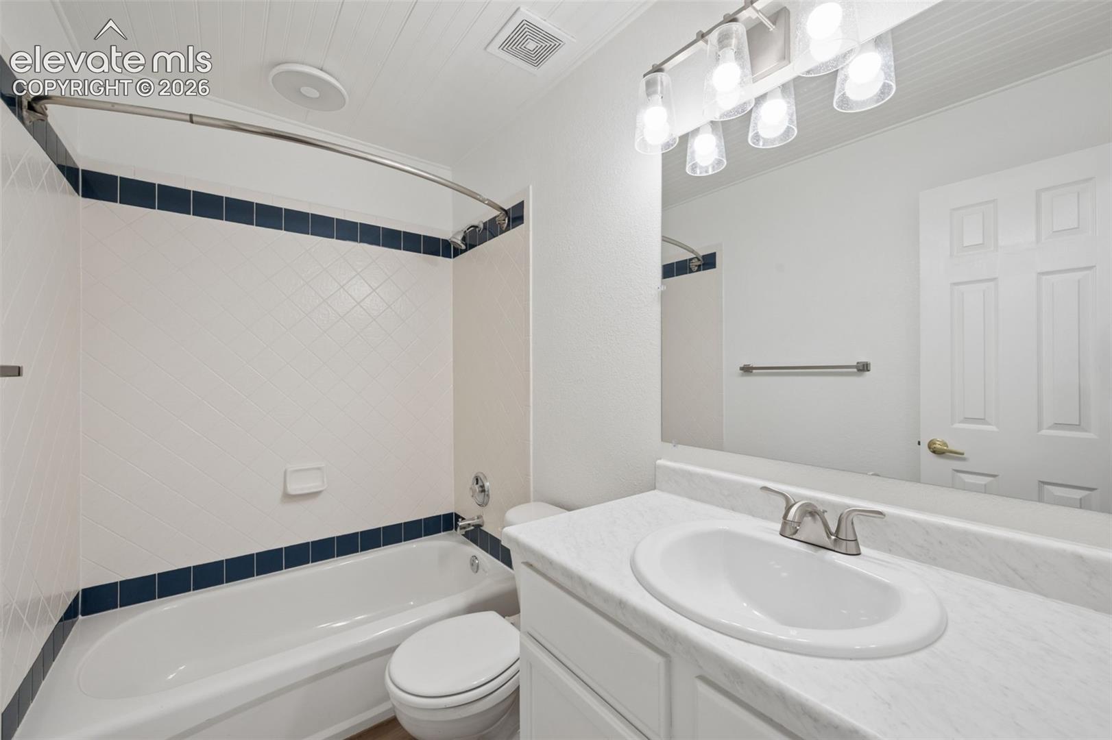 Bathroom featuring white cabinetry, solid-surface countertop with integrated basin, shower-tub combination with decorative accent tile, and multi-bulb vanity lighting