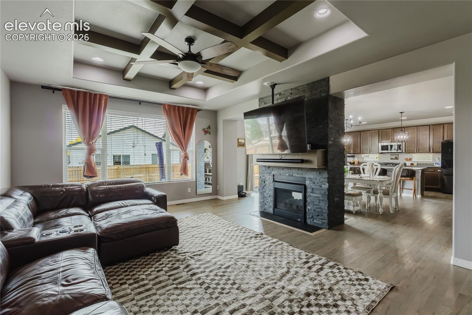 Living area featuring a coffered ceiling with a ceiling fan, a fireplace with stone surround, wood-finish flooring, and a sightline into the kitchen