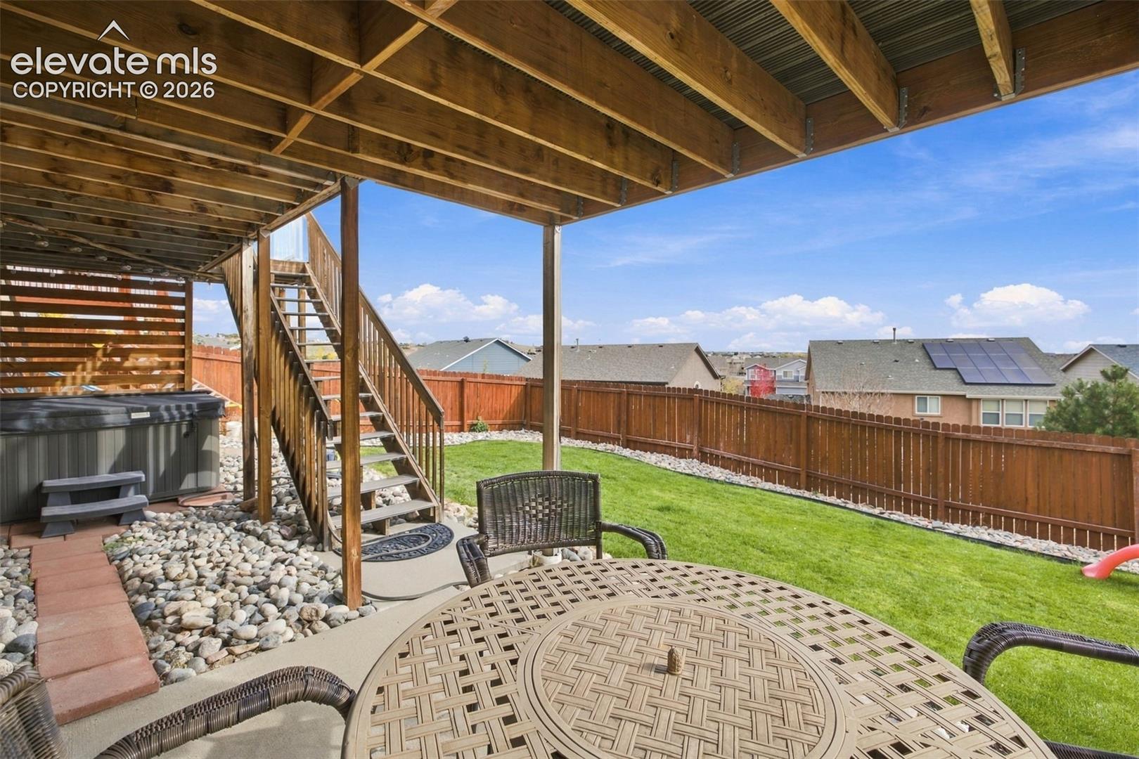Covered concrete patio featuring an exposed timber beam ceiling, wood privacy fencing, a manicured lawn, and a staircase leading to an elevated deck