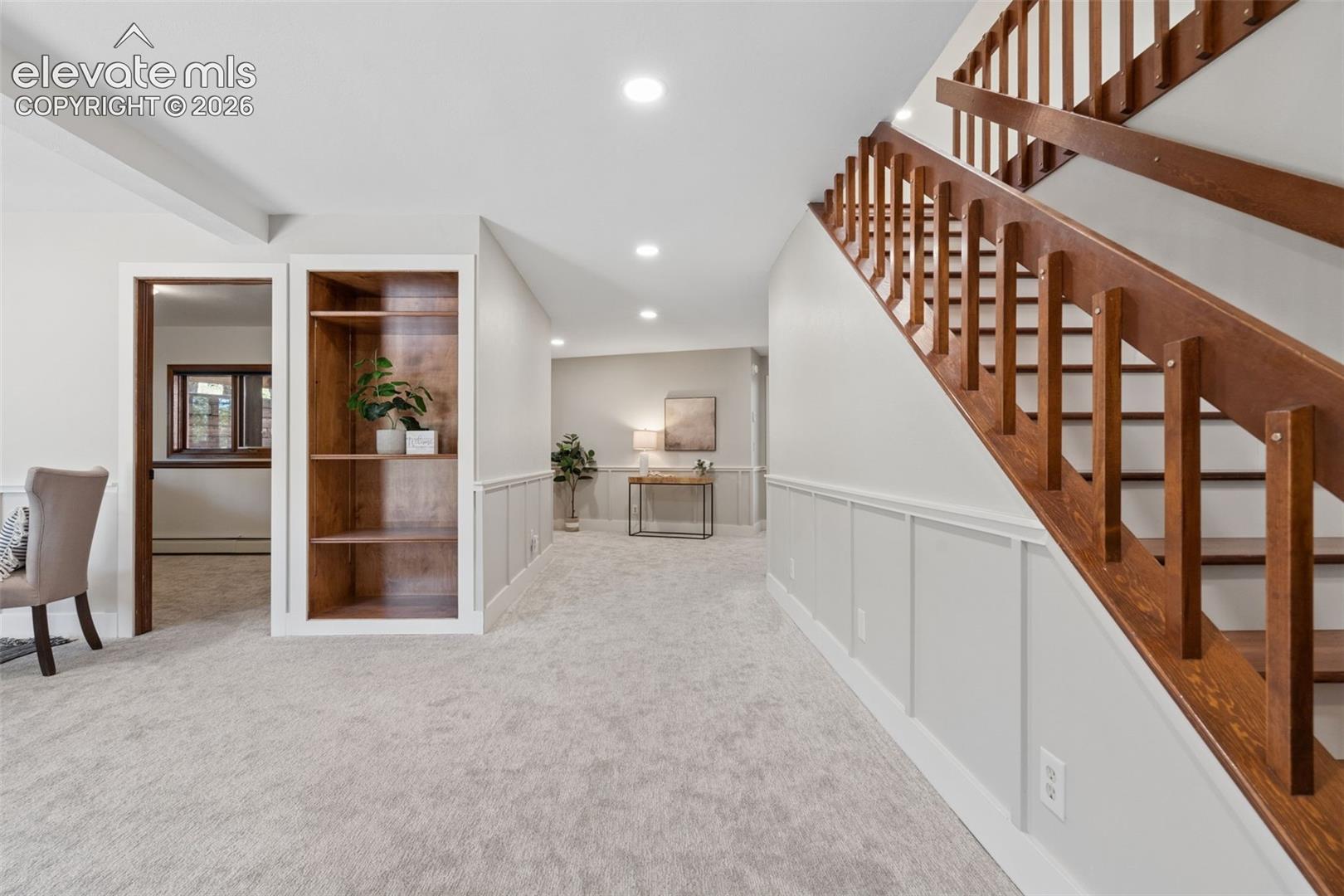 Wood staircase with vertical balusters, light gray wall paneling, built-in wooden shelving, recessed ceiling lighting, and light-toned carpeting