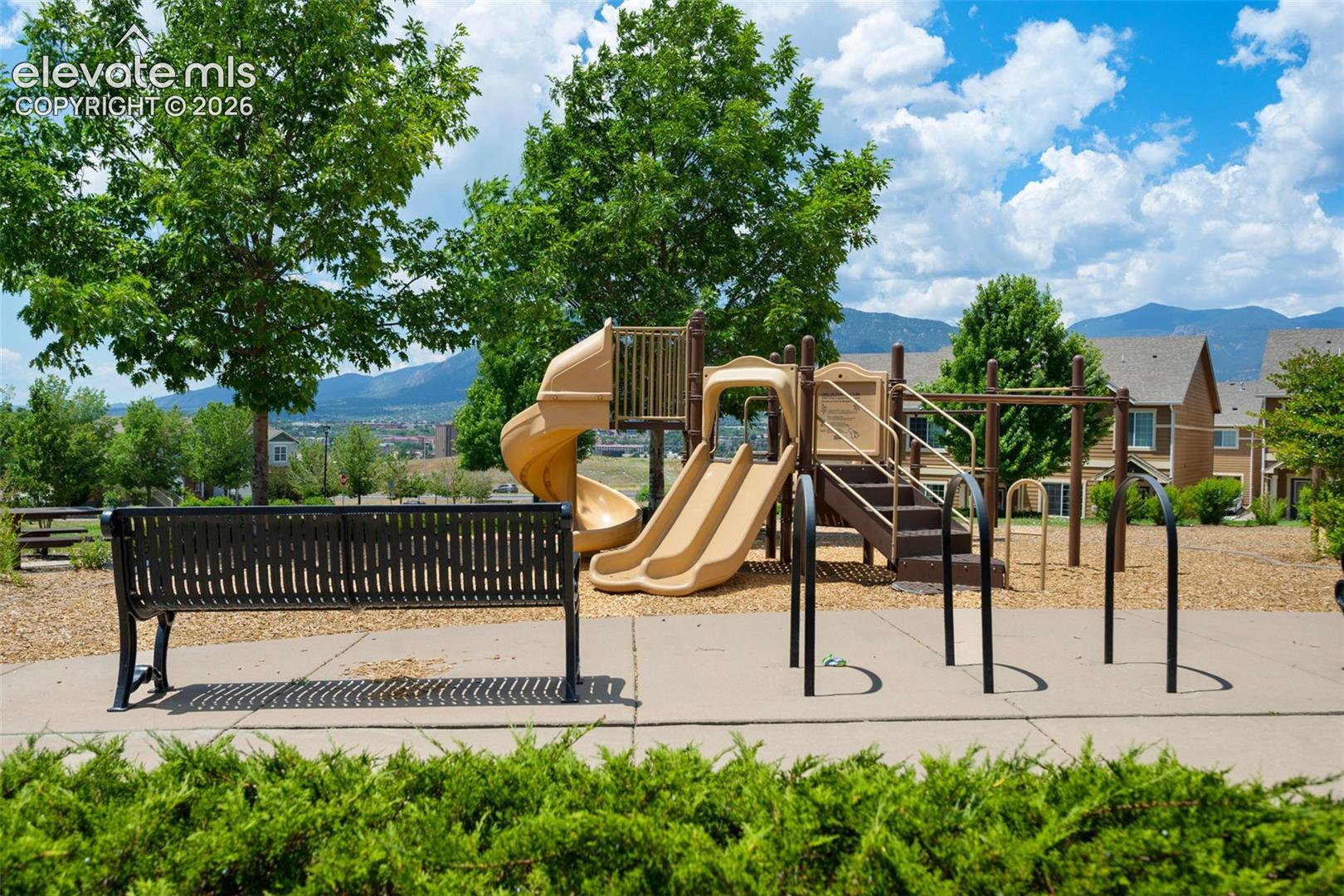 Community playground featuring multiple slides, climbing structures, and swings, set against a backdrop of mature trees and distant mountains