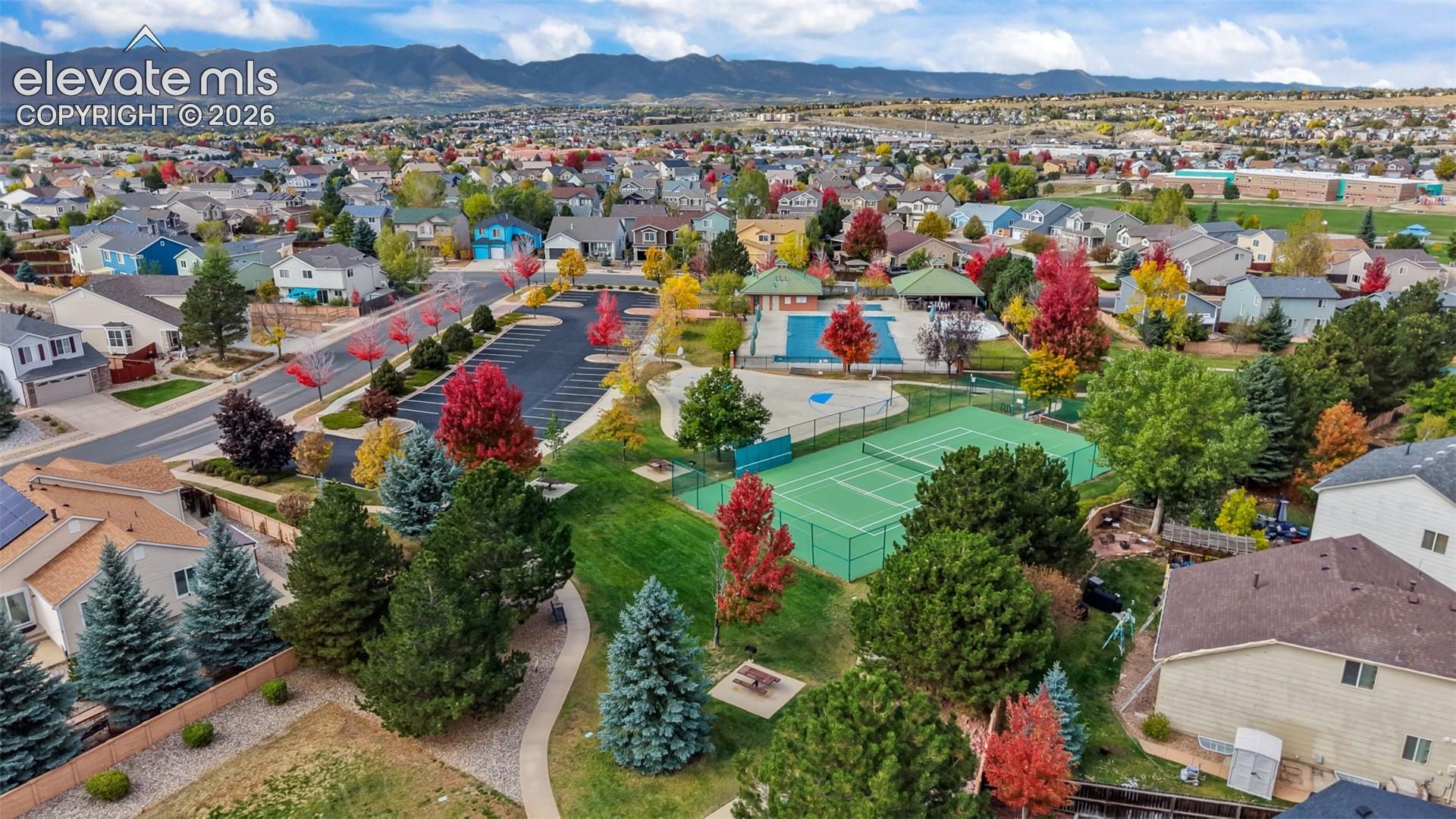 Community recreation area featuring a swimming pool, tennis court, paved pathways, and mature evergreen and deciduous trees against a mountain backdrop