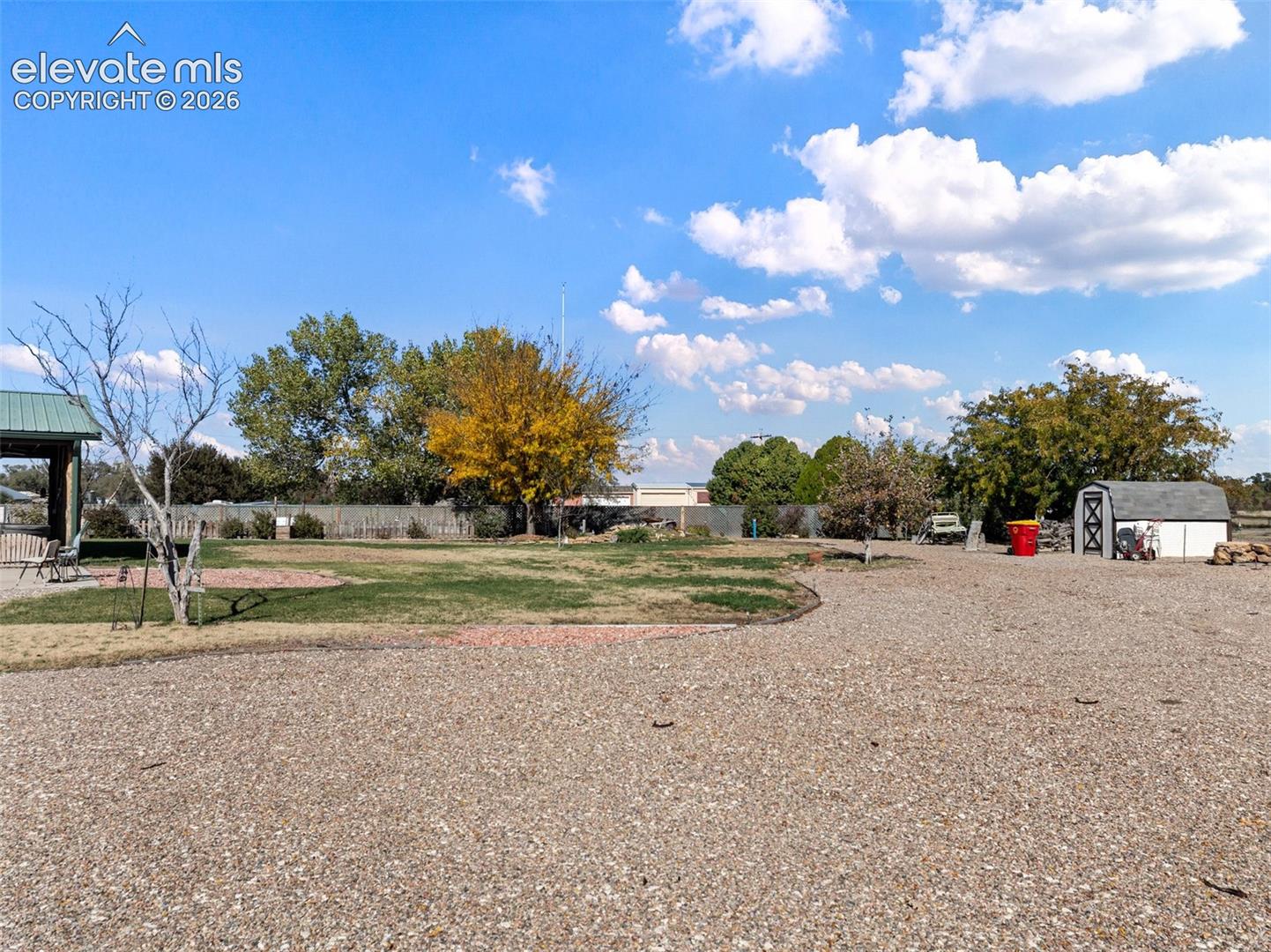 Expansive outdoor space featuring a graveled surface, a covered pavilion structure, a standalone storage shed, and mature trees