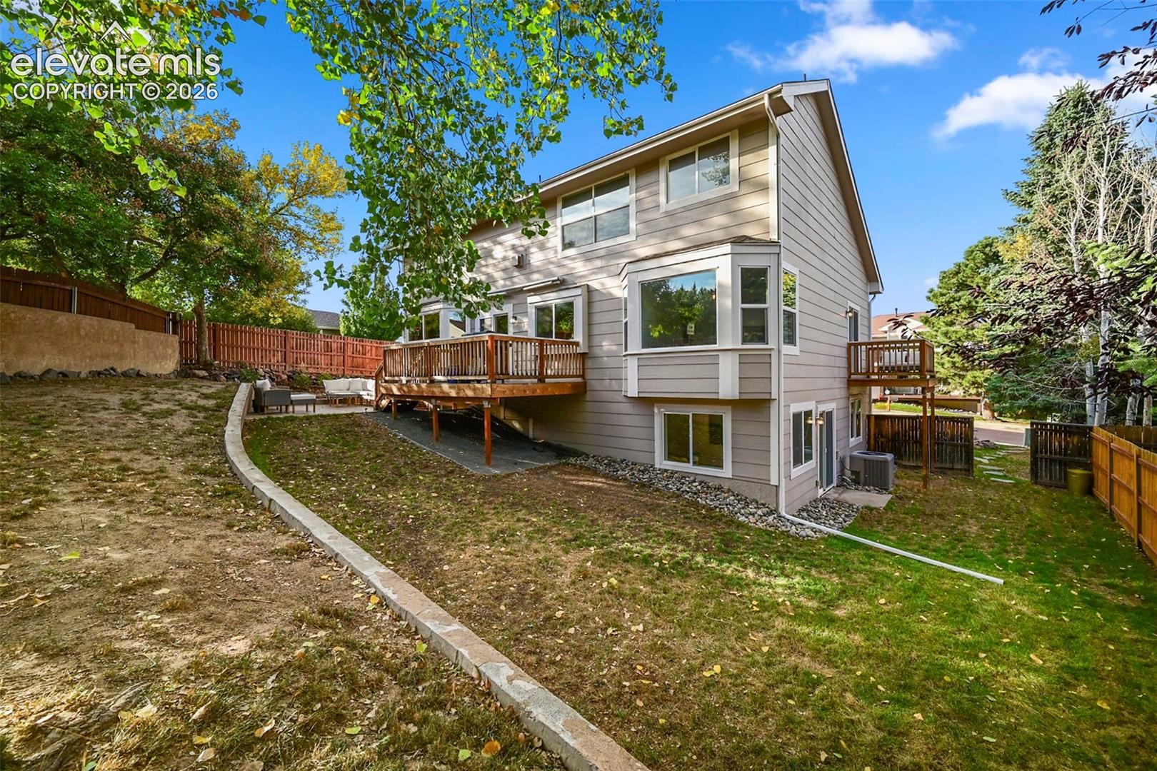Multi-level deck, bay windows, lower level patio access, and a sloped yard with a retaining wall