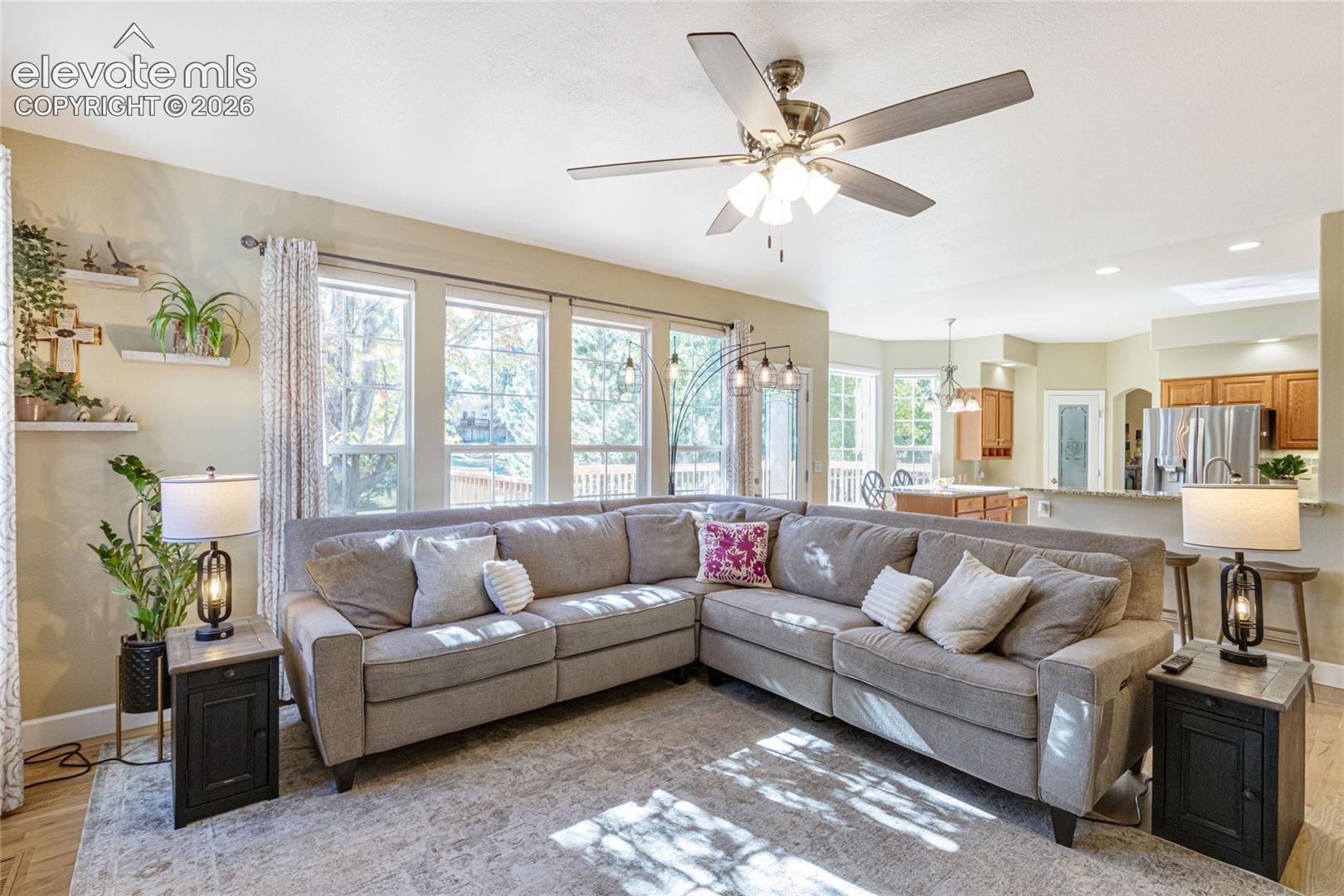 Open-concept living space featuring wood-finish flooring, a multi-blade ceiling fan with integrated lighting, oversized windows, recessed lighting, and a kitchen island with stone-finish countertops