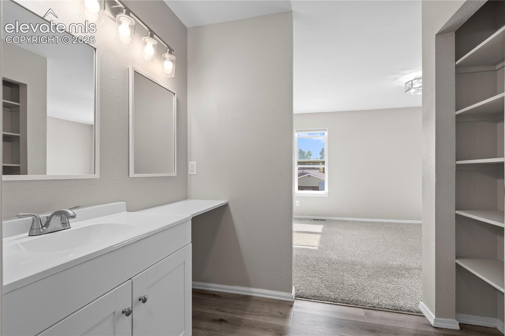 Bathroom vanity featuring a solid-surface countertop, integrated sink, dual rectangular mirrors, and four-bulb overhead lighting fixture, complemented by built-in wall shelving and wood-finish flooring