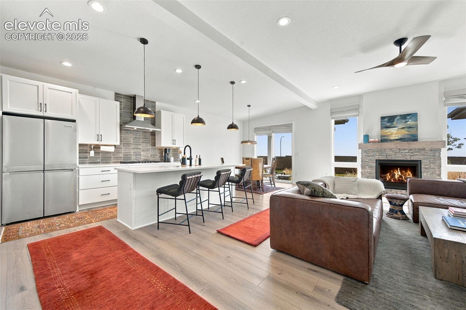 Open-concept kitchen featuring white shaker-style cabinetry, quartz countertops, a stone-tiled backsplash, pendant lighting, and wood-finish flooring with a stone-surround fireplace