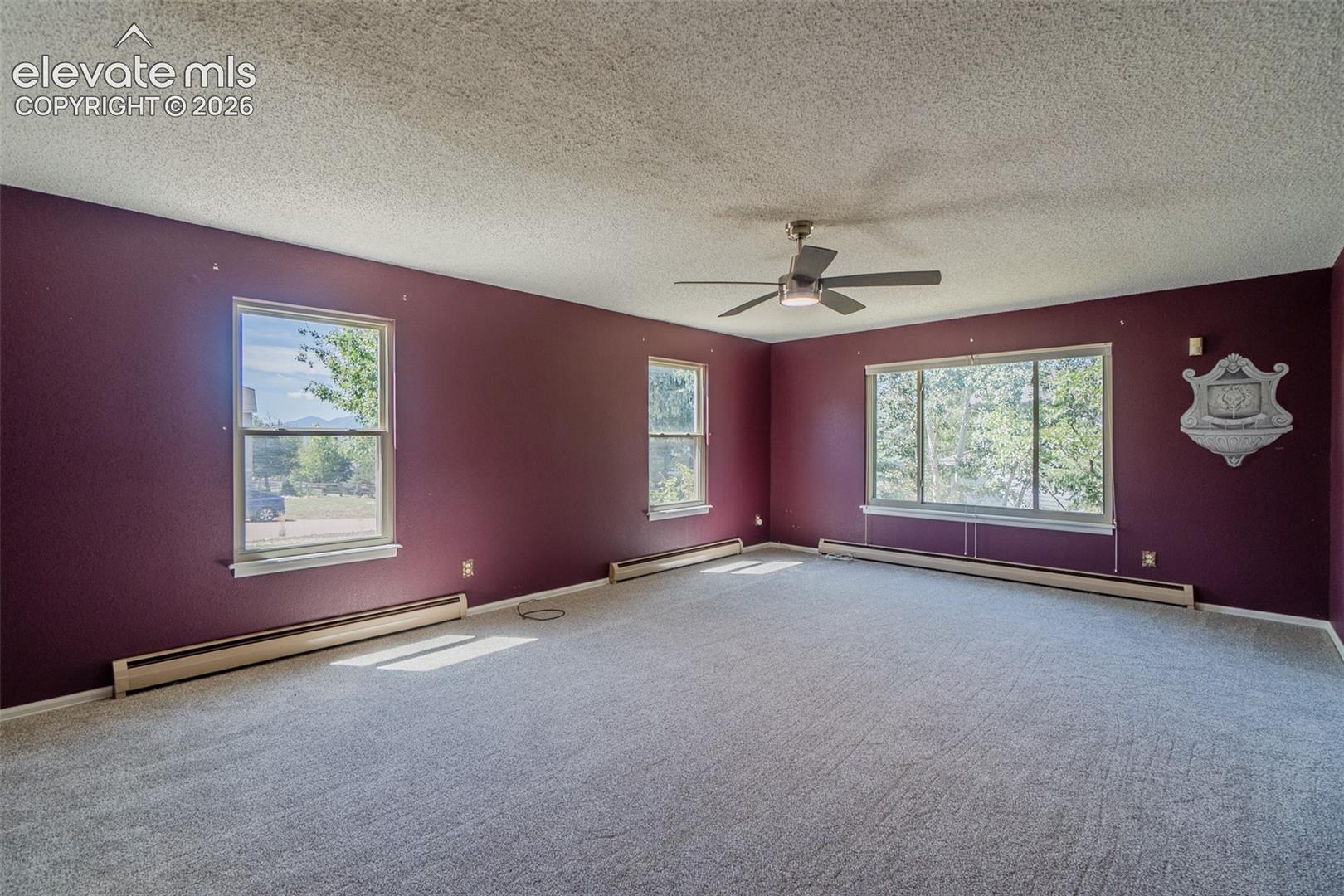 Spacious room with multiple windows, textured ceiling, ceiling fan, and baseboard heating