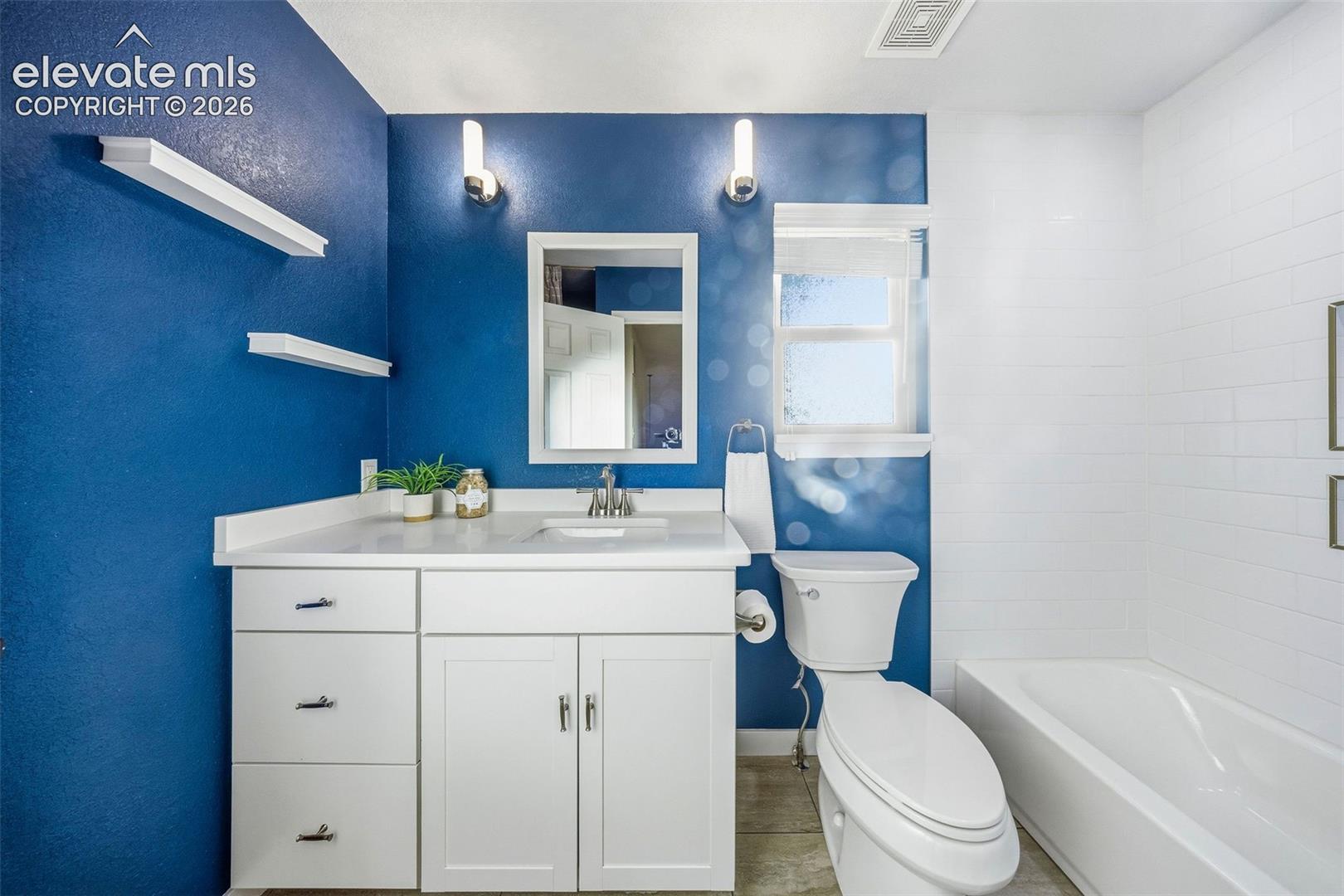 White vanity with countertop basin, wall-mounted light fixtures, built-in floating shelving, and bathtub featuring tiled surround