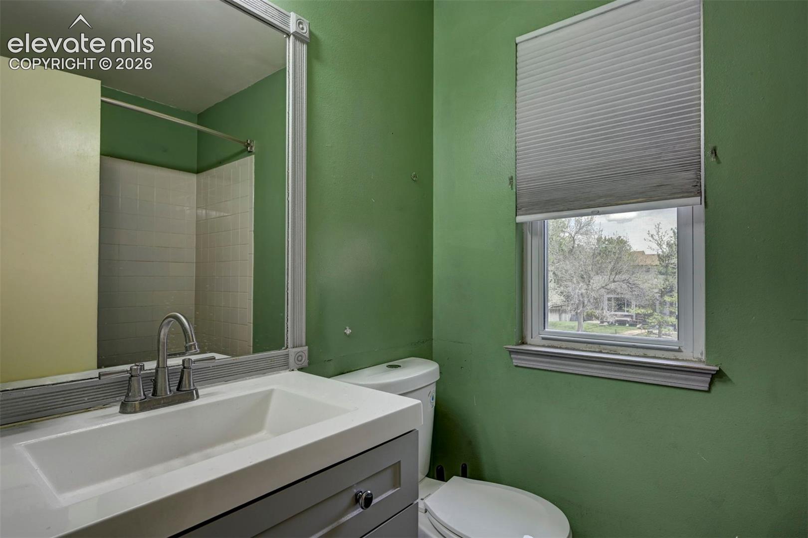 Bathroom featuring a vanity with an integrated sink, brushed nickel faucet, framed mirror, toilet, and a window with pleated shades