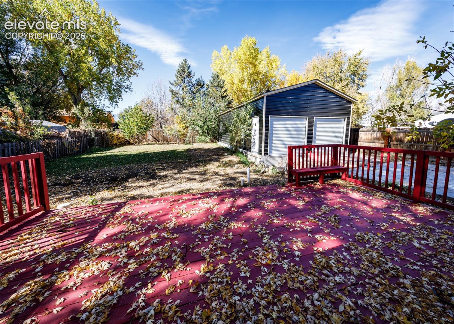 Elevated wood deck featuring red railing and built-in bench seating, large fenced backyard, and a detached two-car garage with white roll-up doors