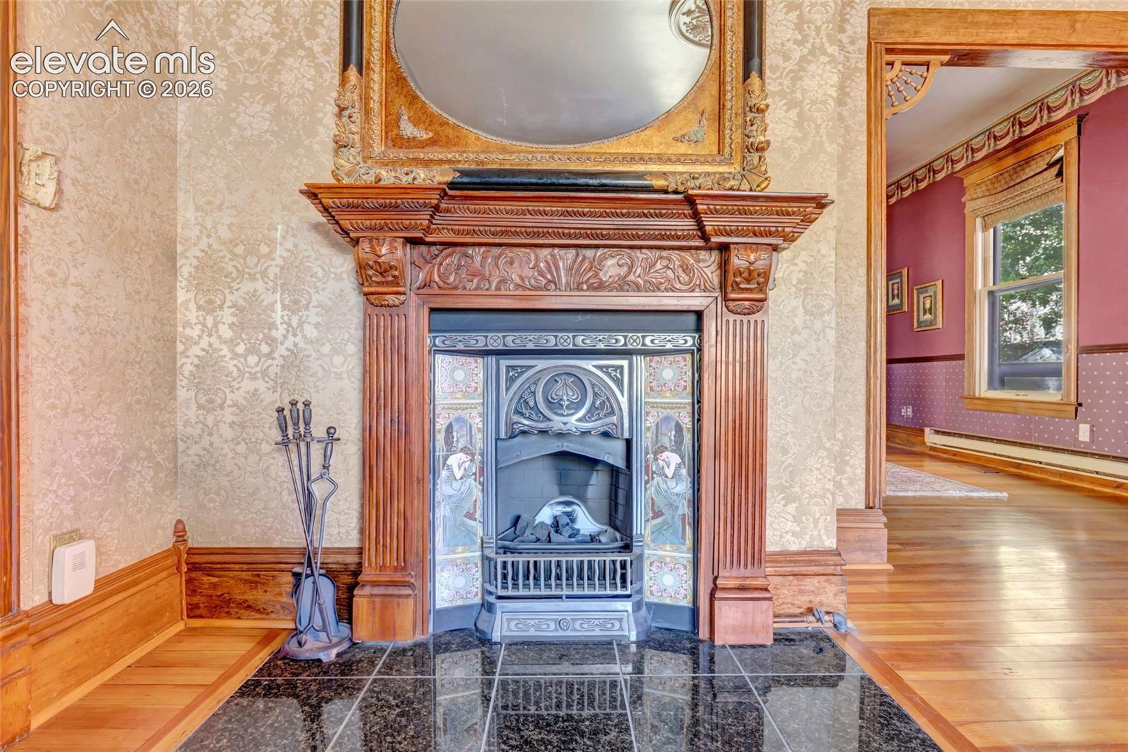 Ornate carved wood fireplace mantel featuring decorative tile surround and metallic firebox insert, set upon a polished stone tile hearth, complemented by wood-finish flooring and detailed wood door casing